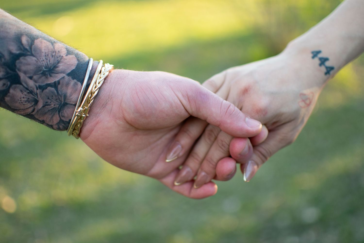 Close-up of a couple holding hands outdoors, showcasing a floral arm tattoo and a 444 wrist tattoo, Grand Island, NE.