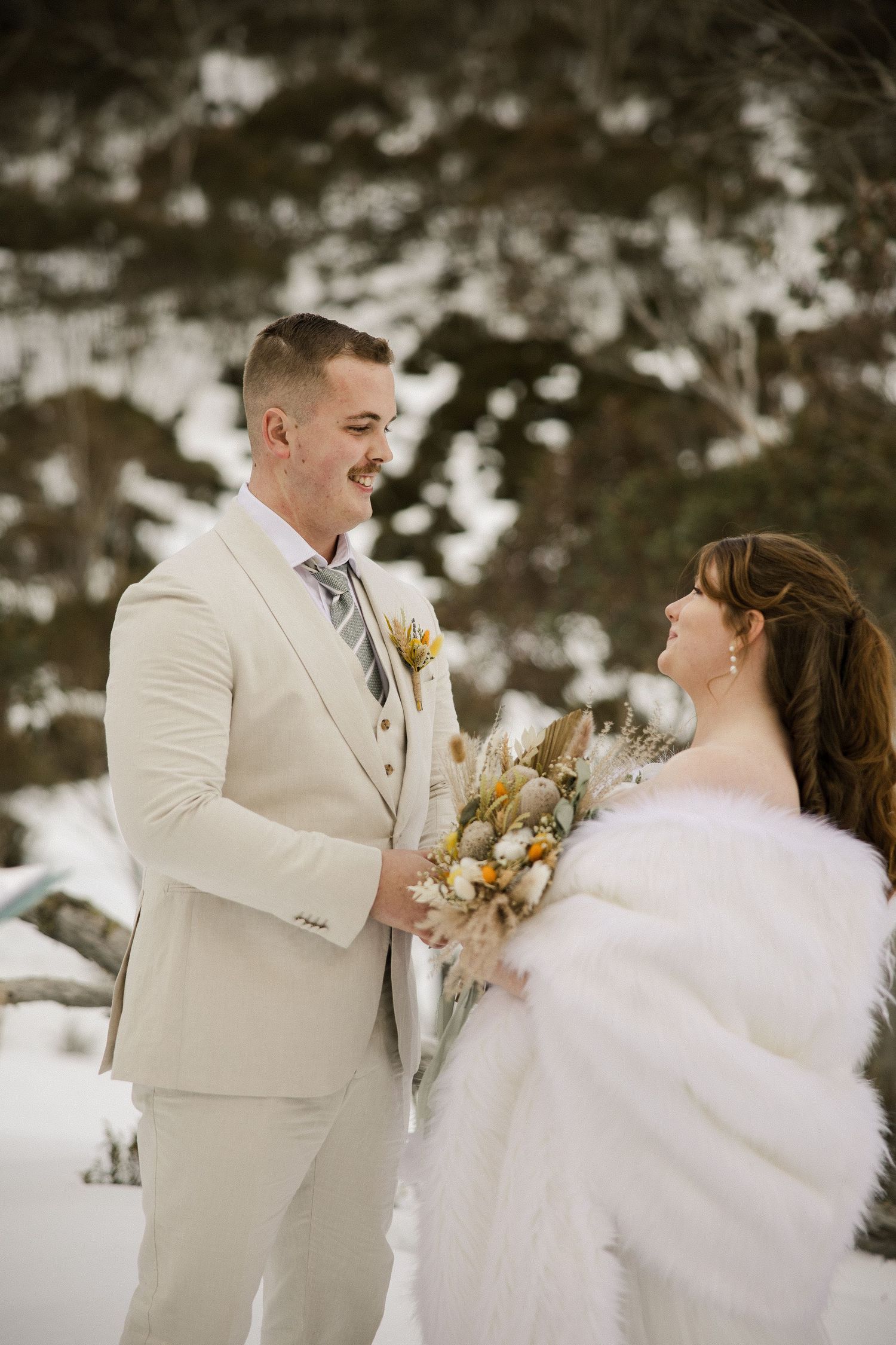 A winter wedding couple shares a joyful moment in the snow, with the bride wearing a white fur wrap and holding dried flowers.