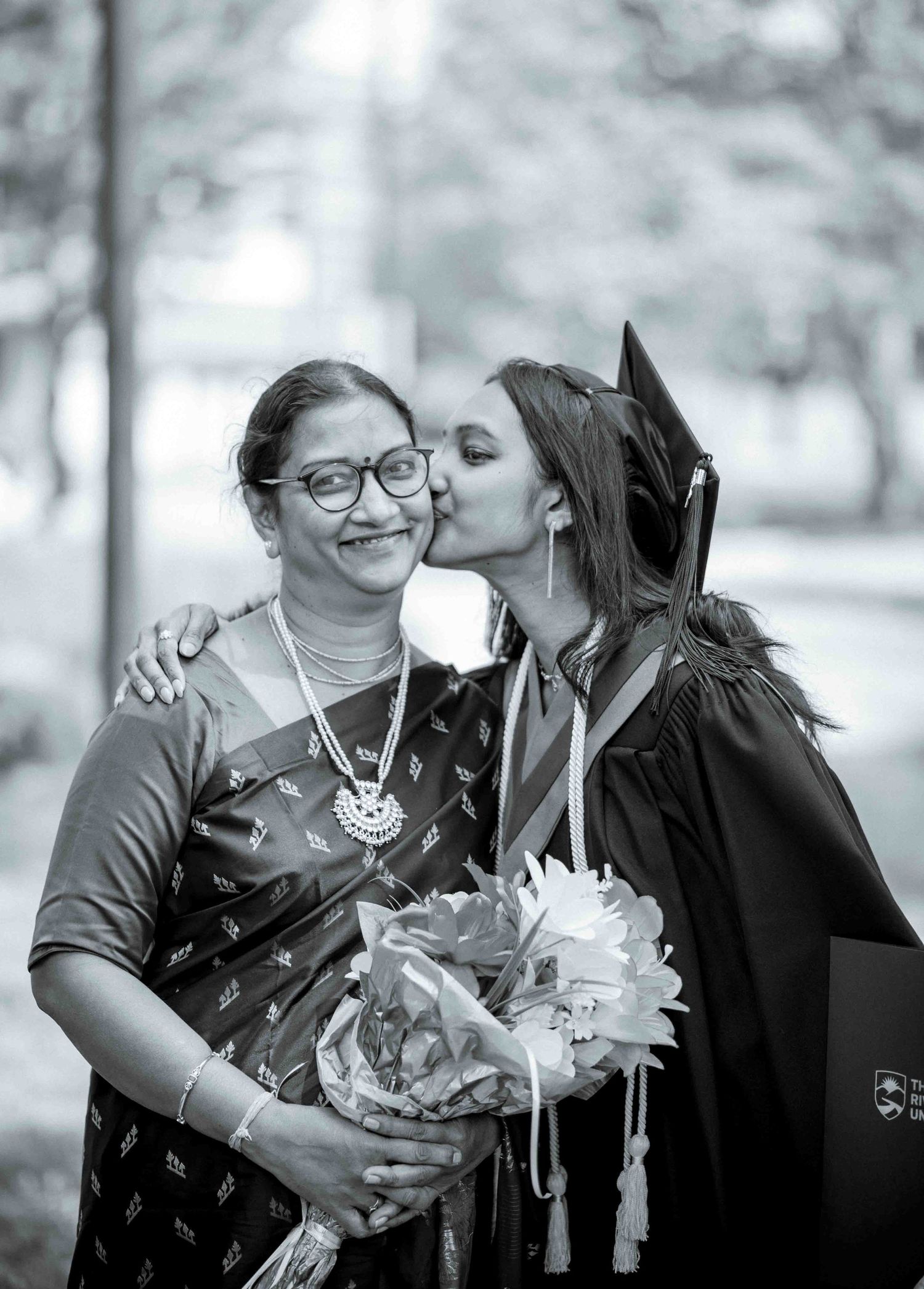Graduate kisses cheek of proud mother holding flowers in black and white graduation photo.