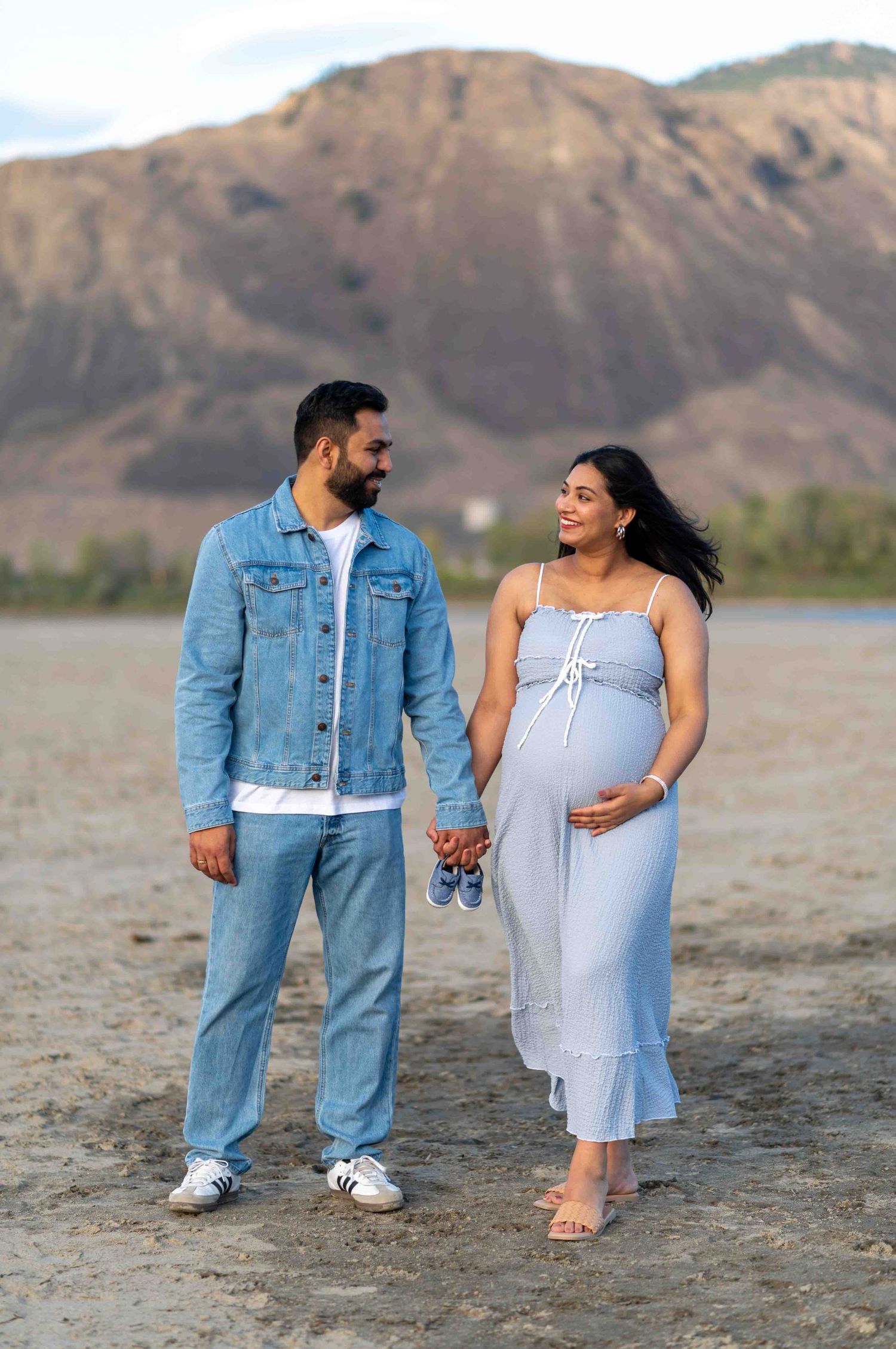 Pregnant couple holding hands on beach with mountain backdrop during maternity photoshoot.