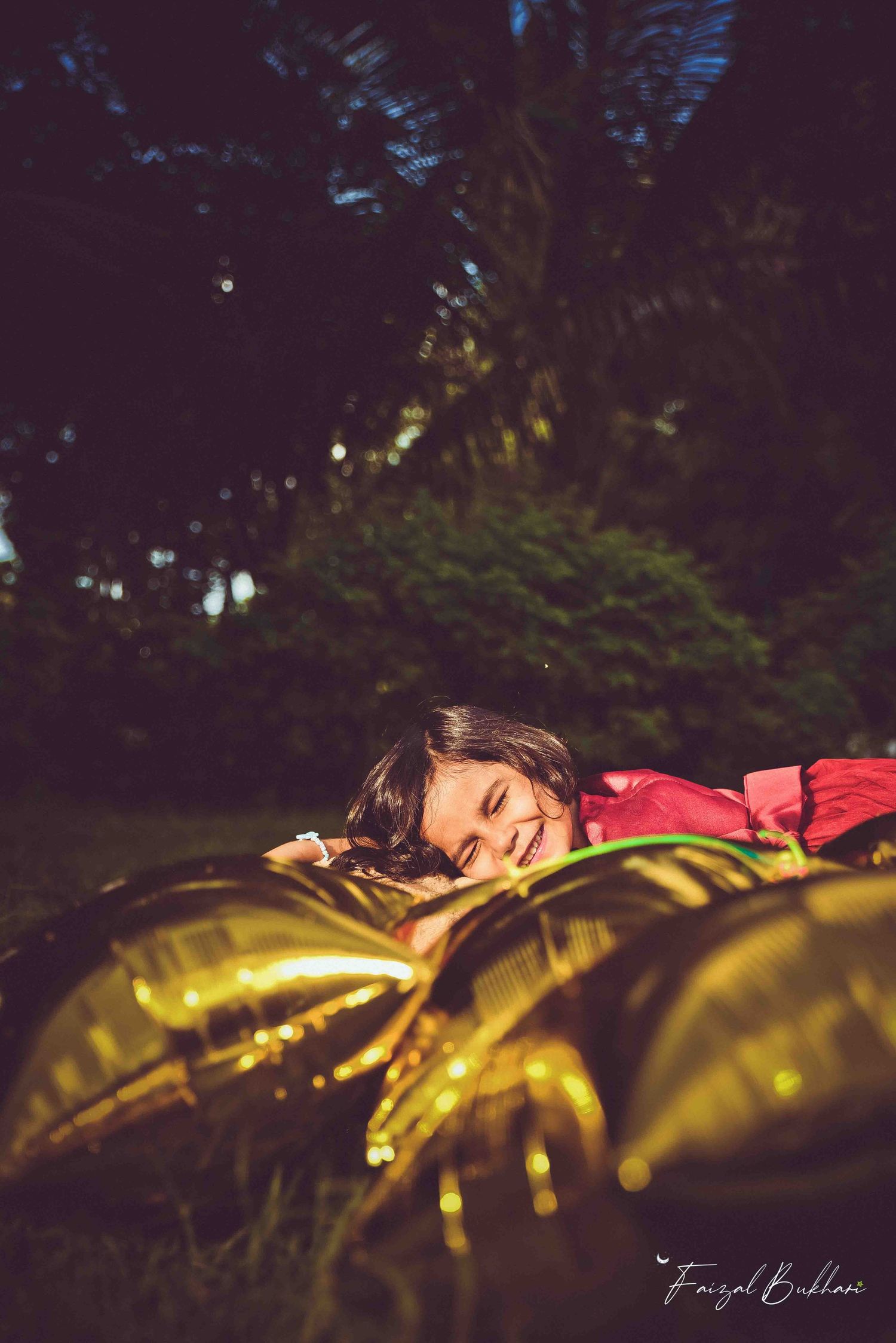 A smiling young girl in red lies on grass beside shiny gold star balloons in a dark garden.