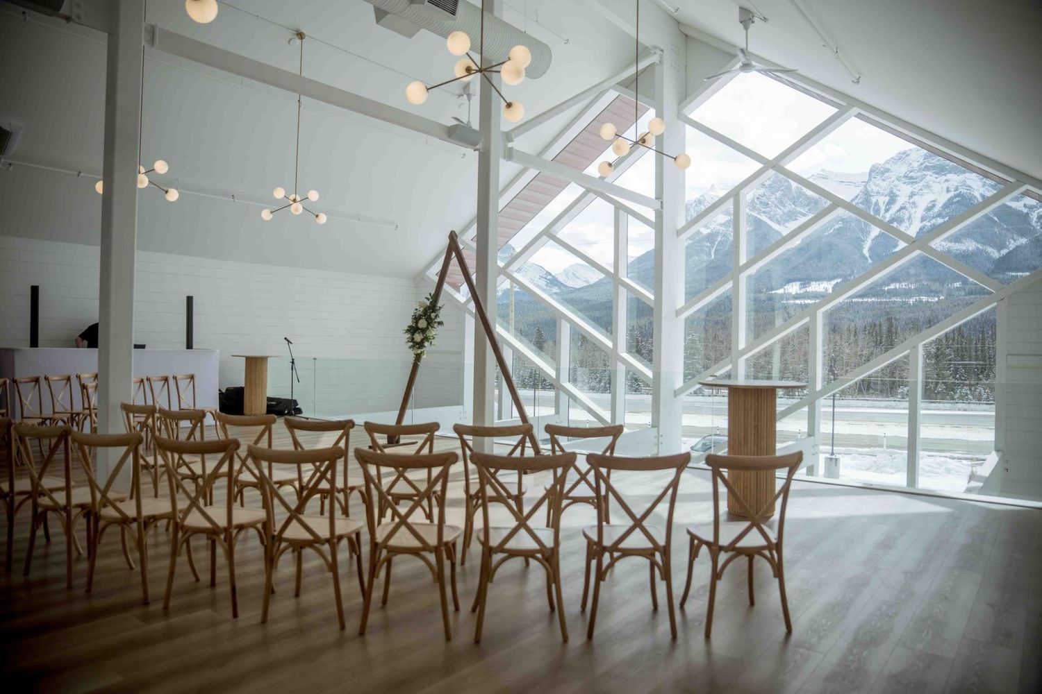 Elegant wedding ceremony venue with wooden chairs, triangular arch, and stunning mountain views through floor-to-ceiling windows.