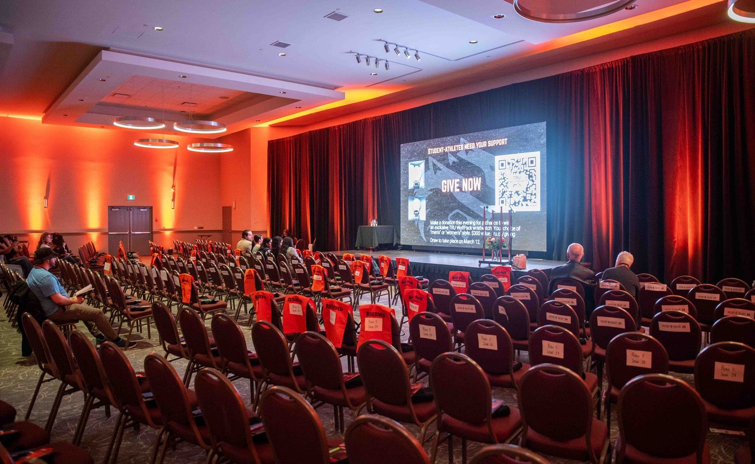 Spacious conference ballroom with rows of red chairs facing a stage with a fundraising donation QR code displayed.