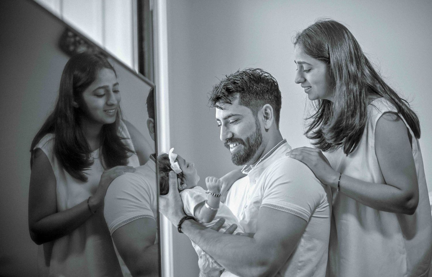 Black and white photo of man holding newborn baby with two women looking on near mirror.