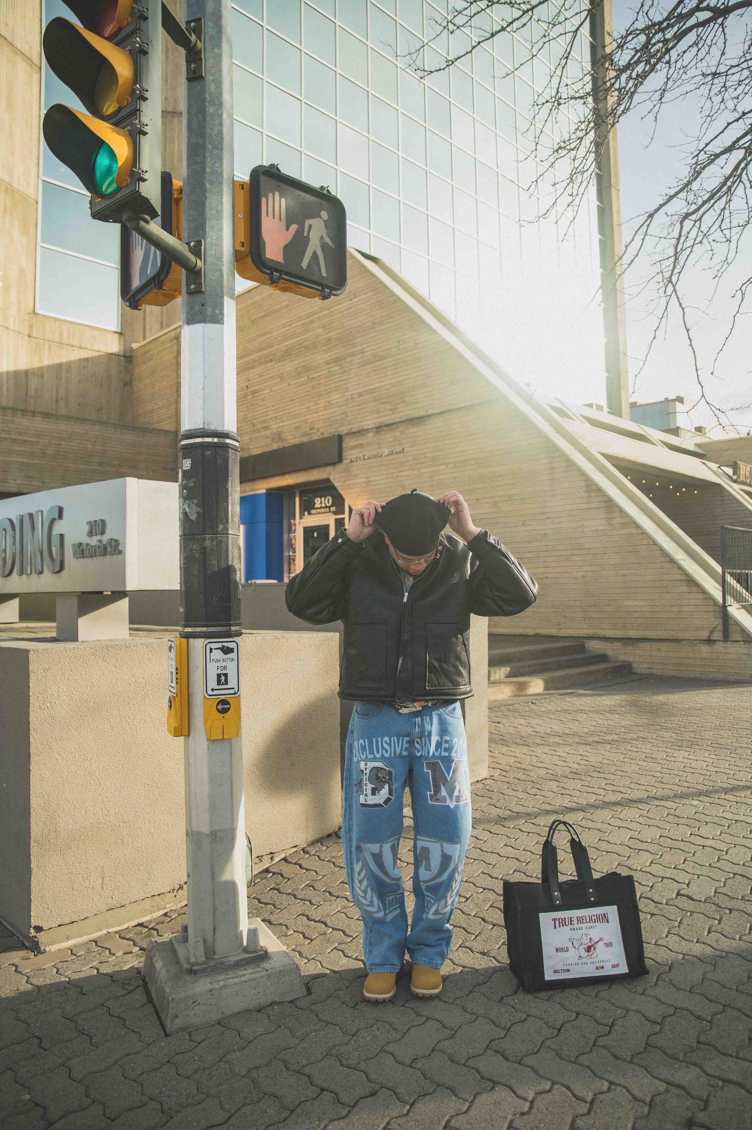 Man adjusting cap at sunny city intersection wearing blue graphic jeans beside True Religion tote bag.