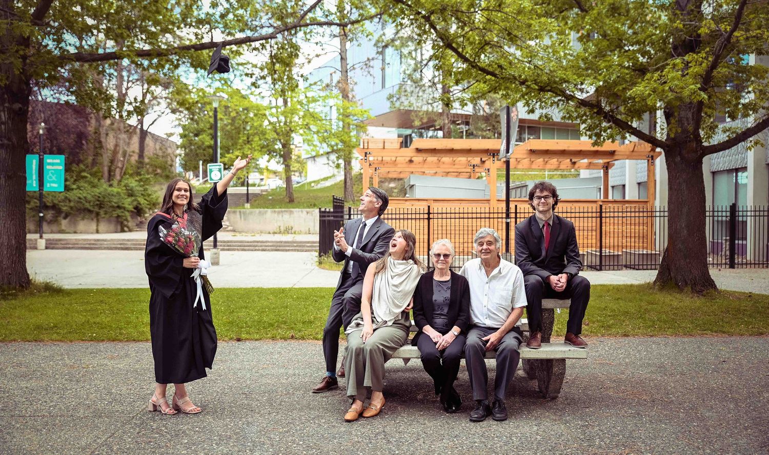 Graduate in black gown celebrating with family group seated and standing under tree on university campus.