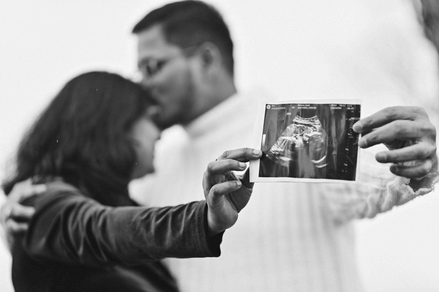 Couple kissing while holding ultrasound sonogram photo, black and white pregnancy announcement portrait.