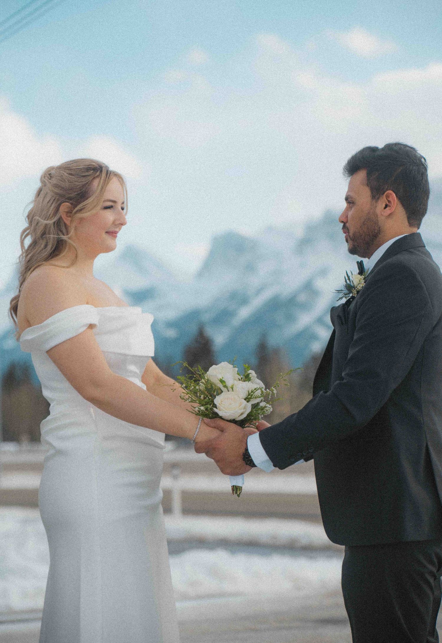 Bride in white off-shoulder gown and groom holding hands with white rose bouquet and snowy mountains.