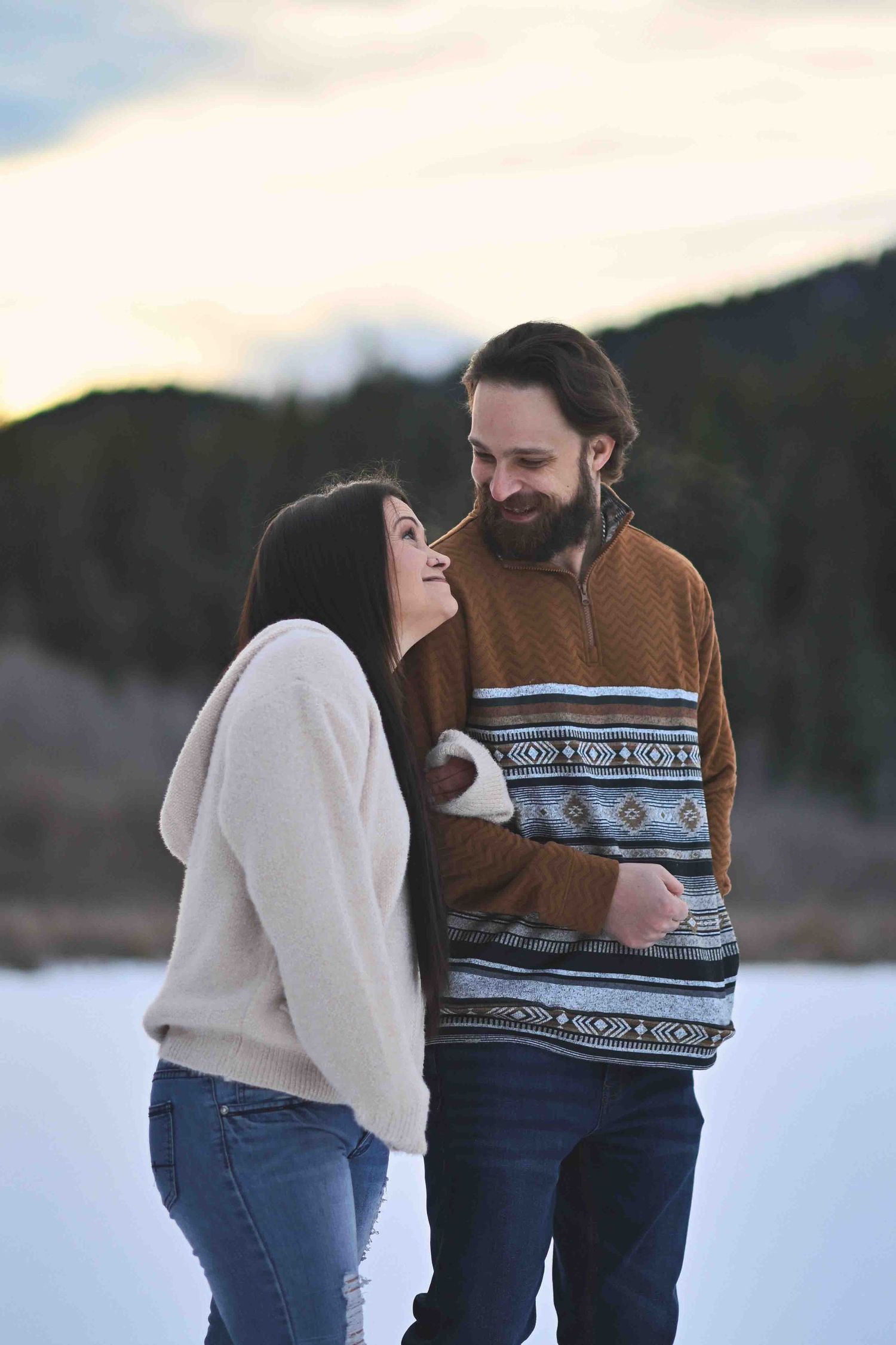 Happy couple laughing together in a snowy winter landscape at dusk during engagement photo session.