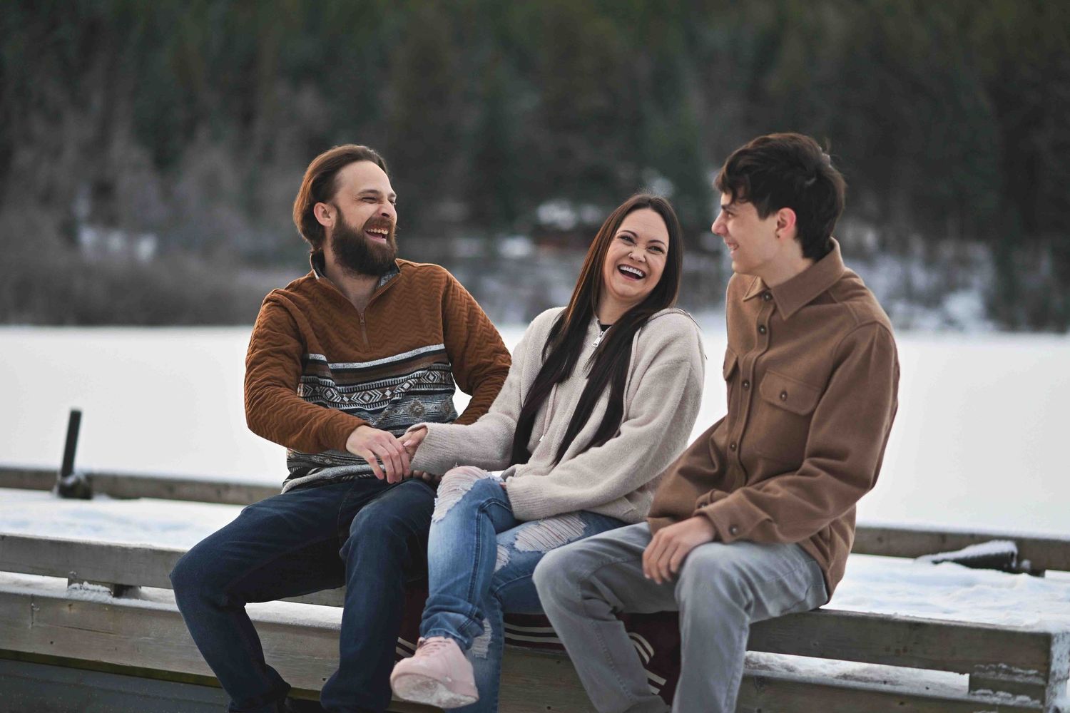 Three friends sitting on a snowy dock laughing together during a fun winter outdoor photo session.