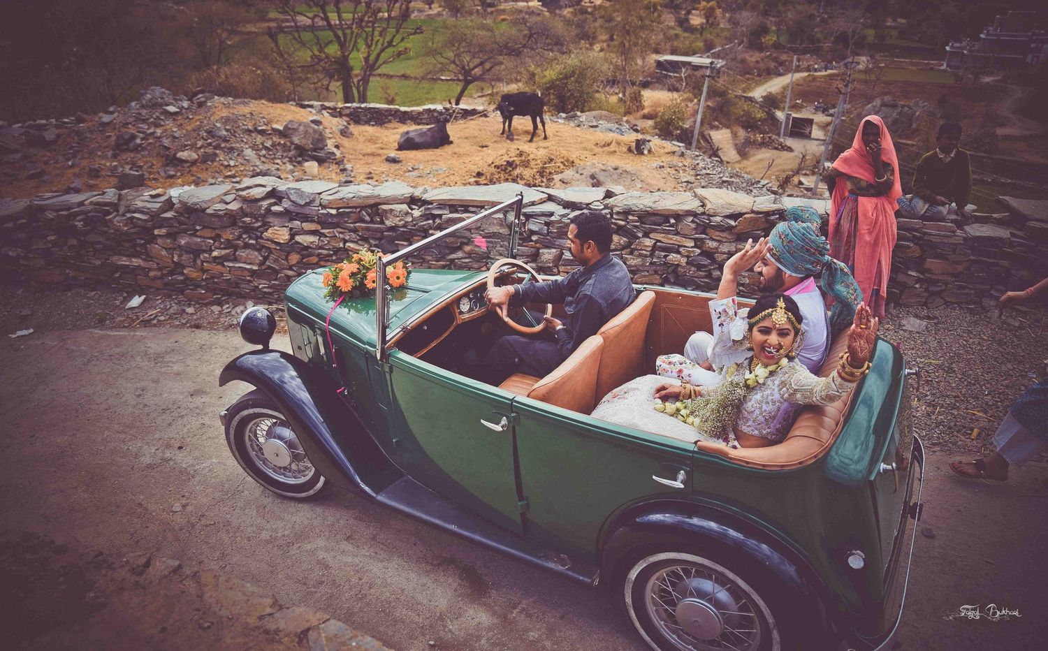 Bride and groom riding in a vintage green convertible car through a rustic Indian countryside landscape.