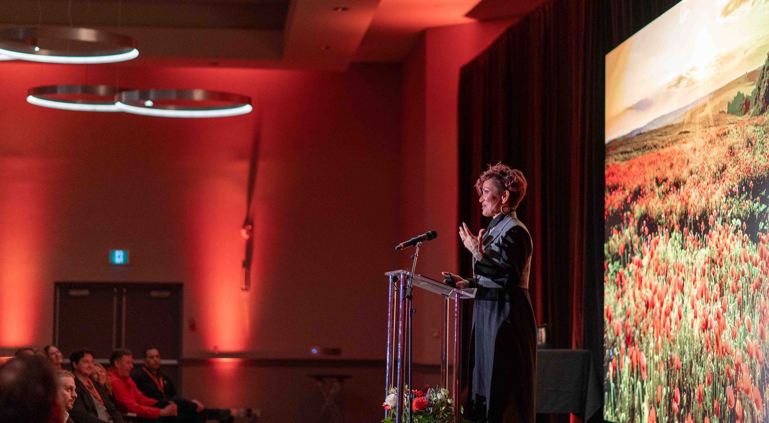 A woman speaks at a glass podium on a red-lit stage with a floral landscape projected behind her.