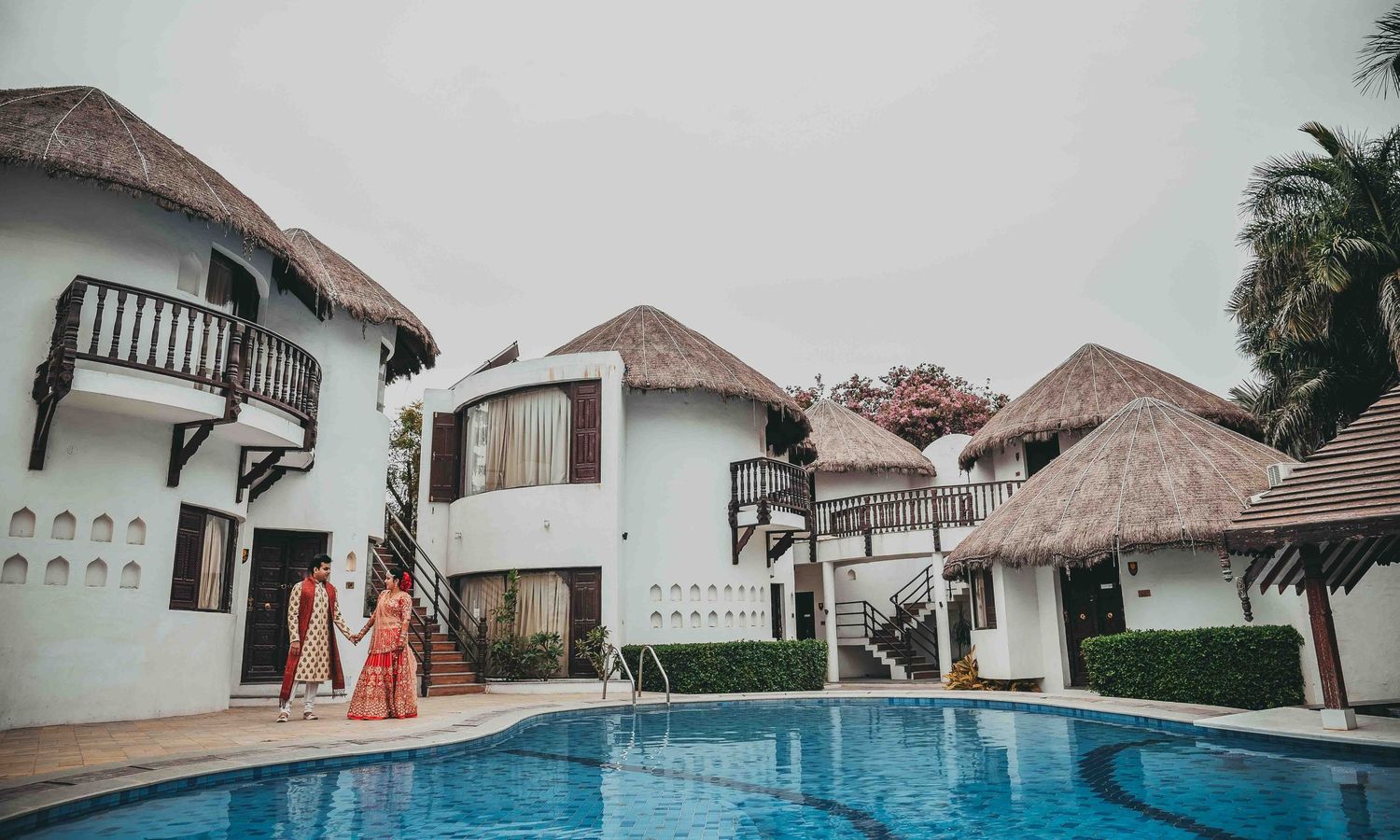 Indian wedding couple in traditional attire posing near a resort pool surrounded by white villas and thatched roofs.