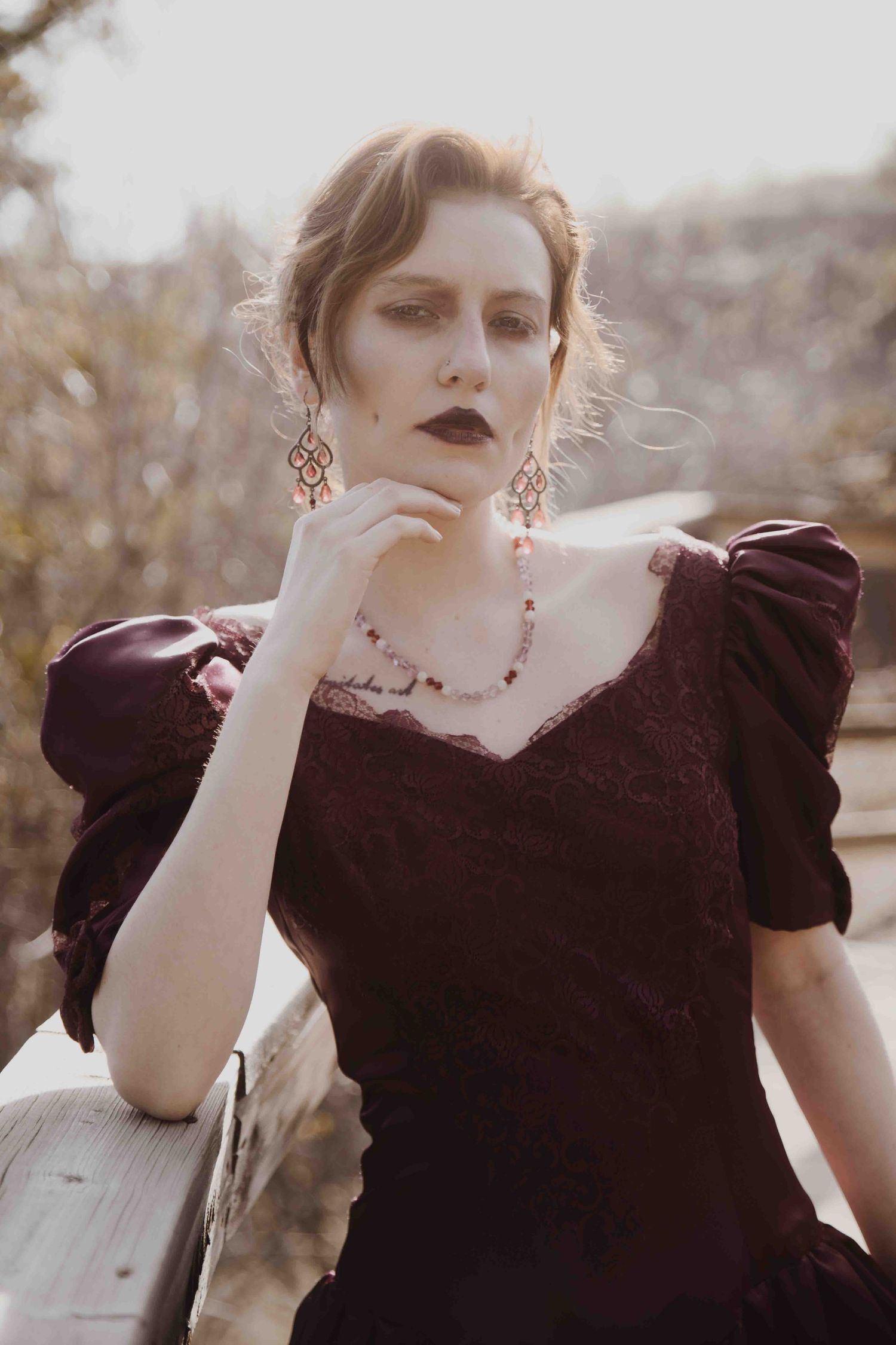 Woman in dark burgundy Victorian lace dress with ornate earrings poses thoughtfully on rustic wooden fence.
