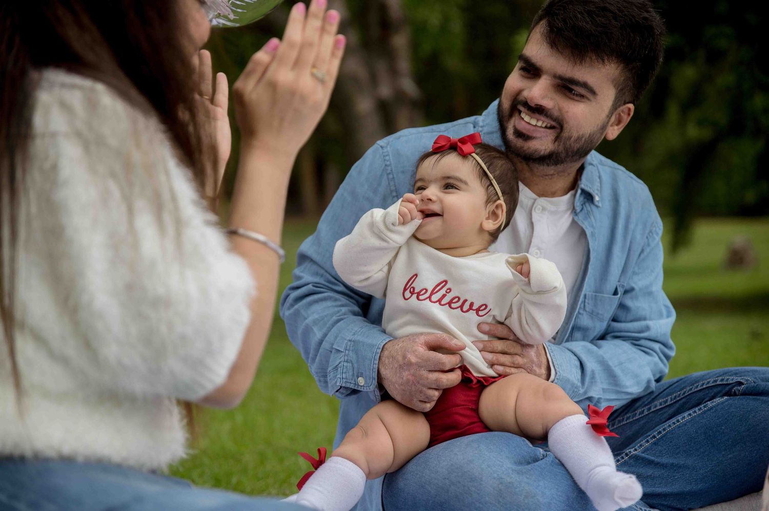 Father holding baby girl with believe pillow while mother claps outdoors on grass.
