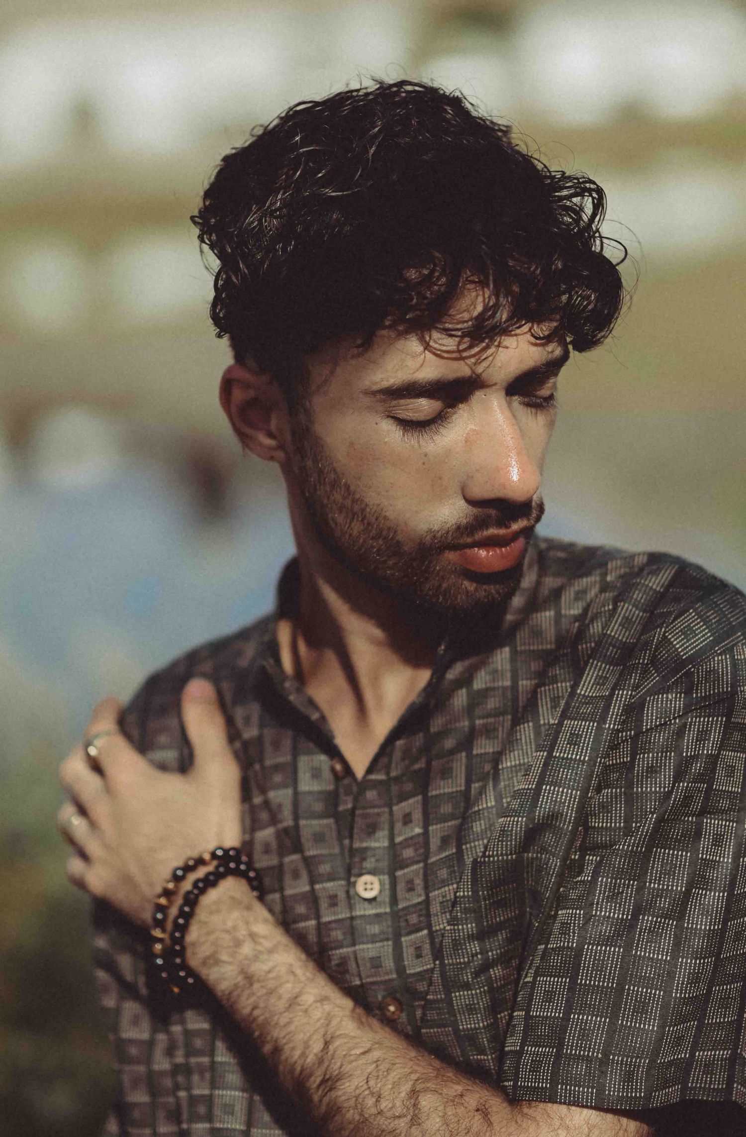 A bearded man with curly hair and a beaded bracelet poses with eyes closed in warm sunlight.