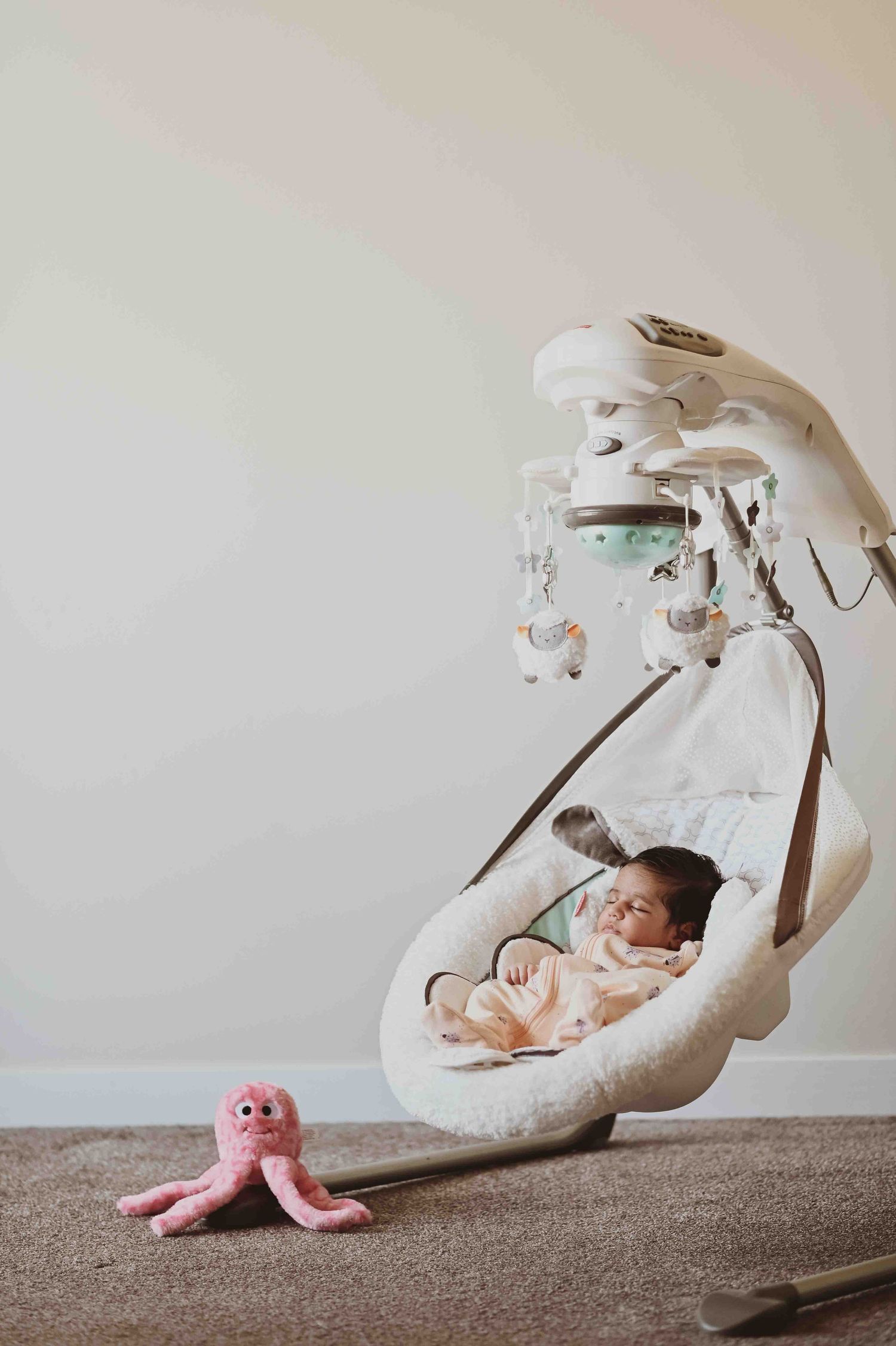 Newborn baby rests in a white fluffy baby swing with a pink octopus toy nearby.