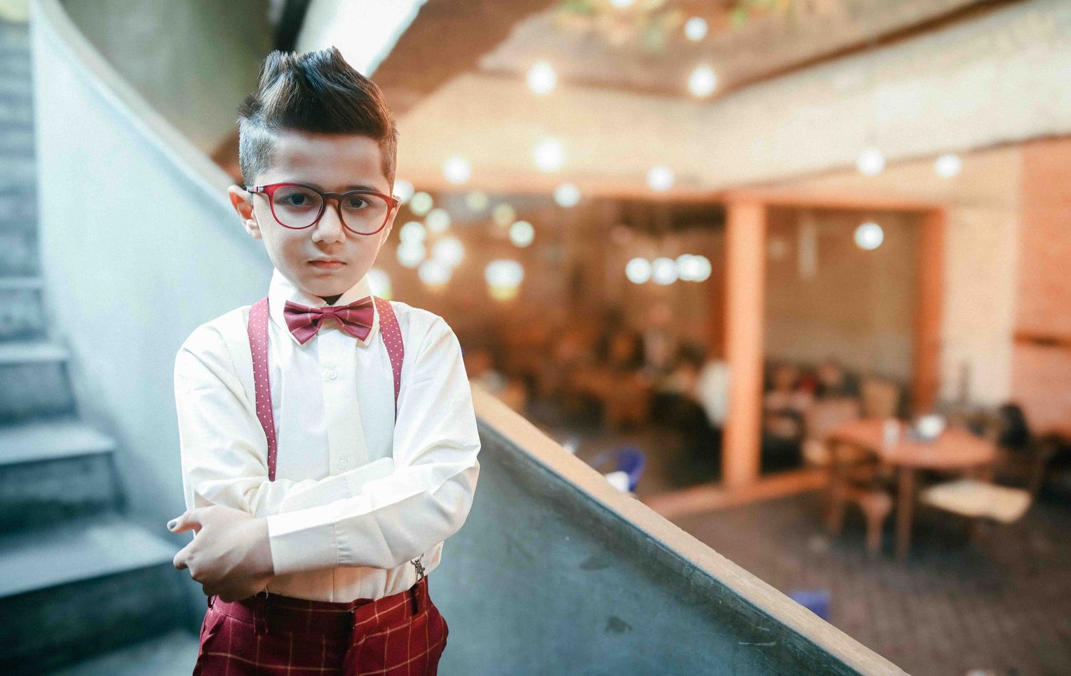 Confident boy in glasses, bow tie and suspenders posing with arms crossed in indoor setting.