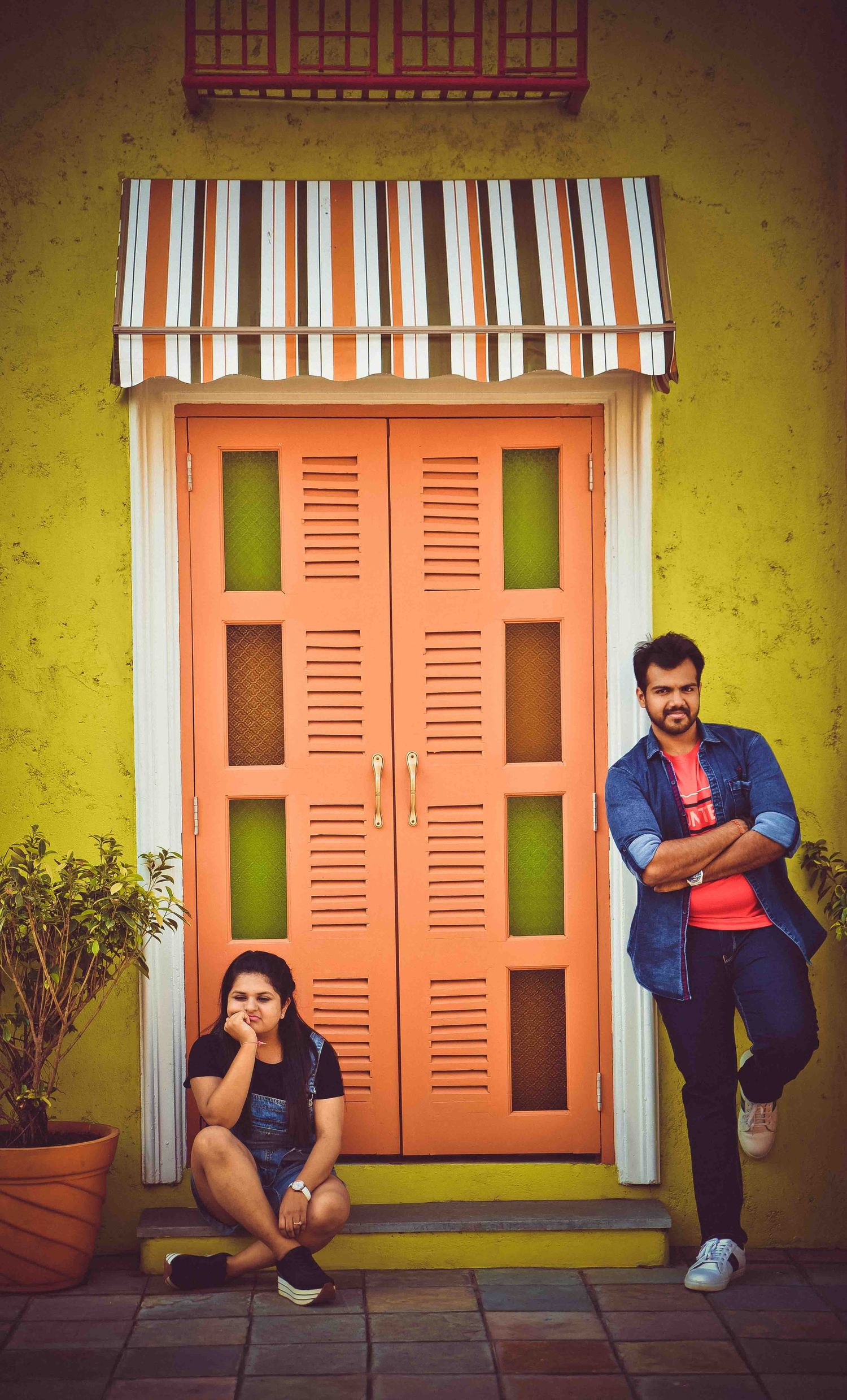 A cheerful Indian couple poses playfully outside a colorful yellow building with an orange door during a pre-wedding shoot.