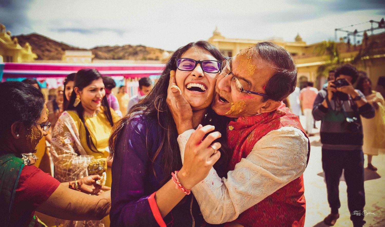Joyful Indian family celebrating Holi with laughter and colorful powder at a vibrant outdoor wedding event.