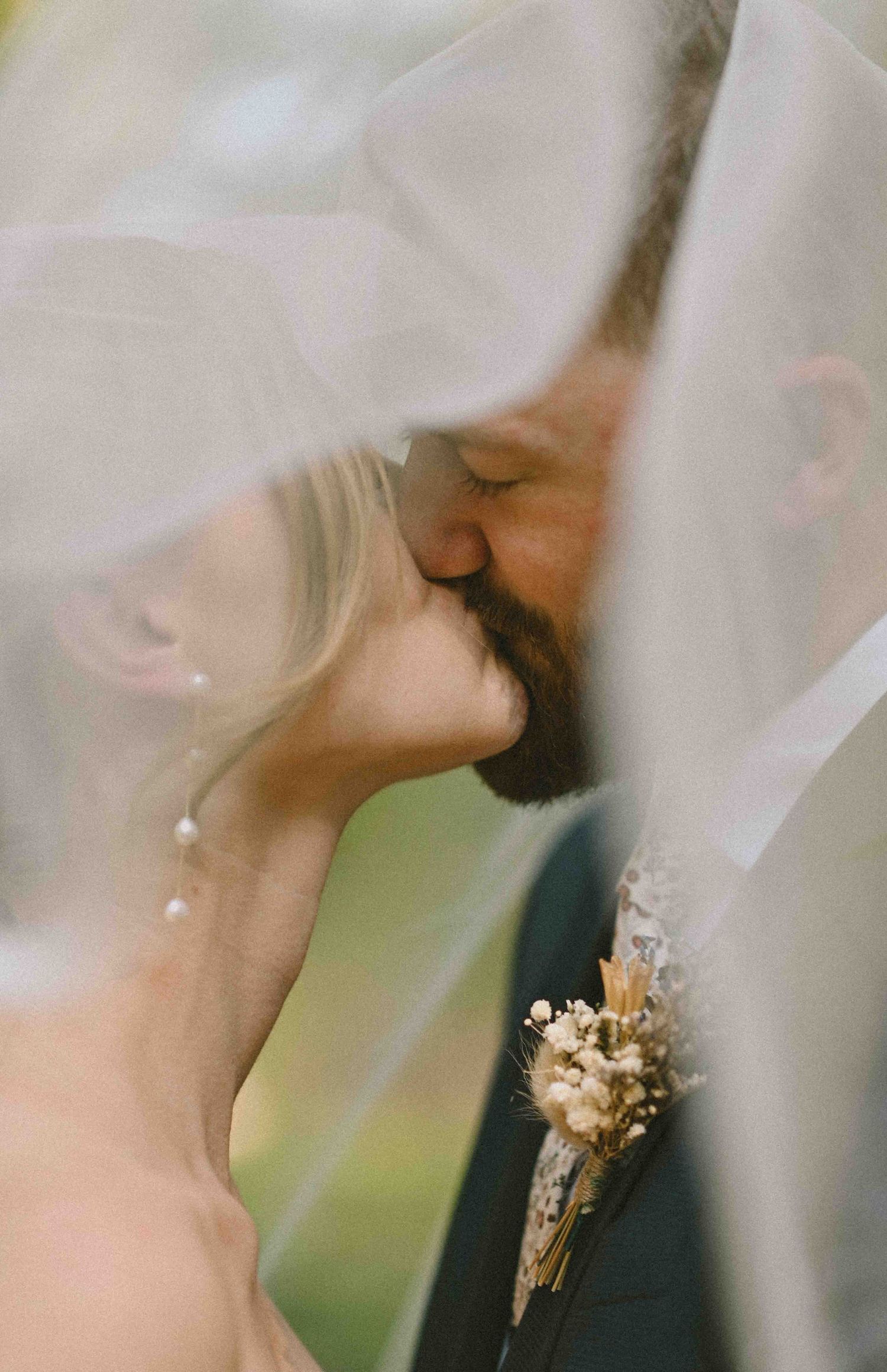 Romantic close-up of bride and groom kissing under flowing white veil, groom wearing dried flower boutonniere.