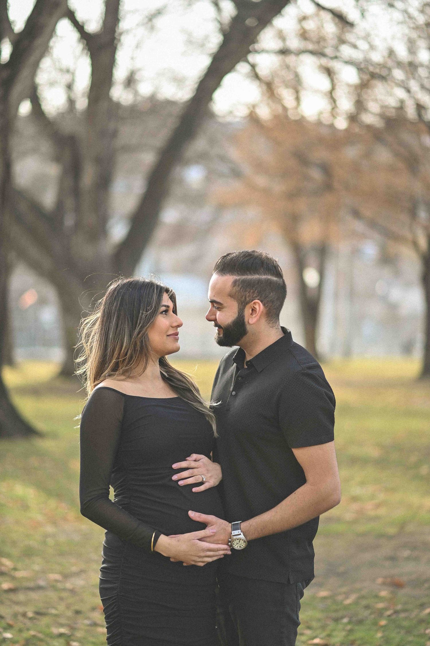 Couple in black outfits sharing a tender maternity moment in a park with autumn trees.
