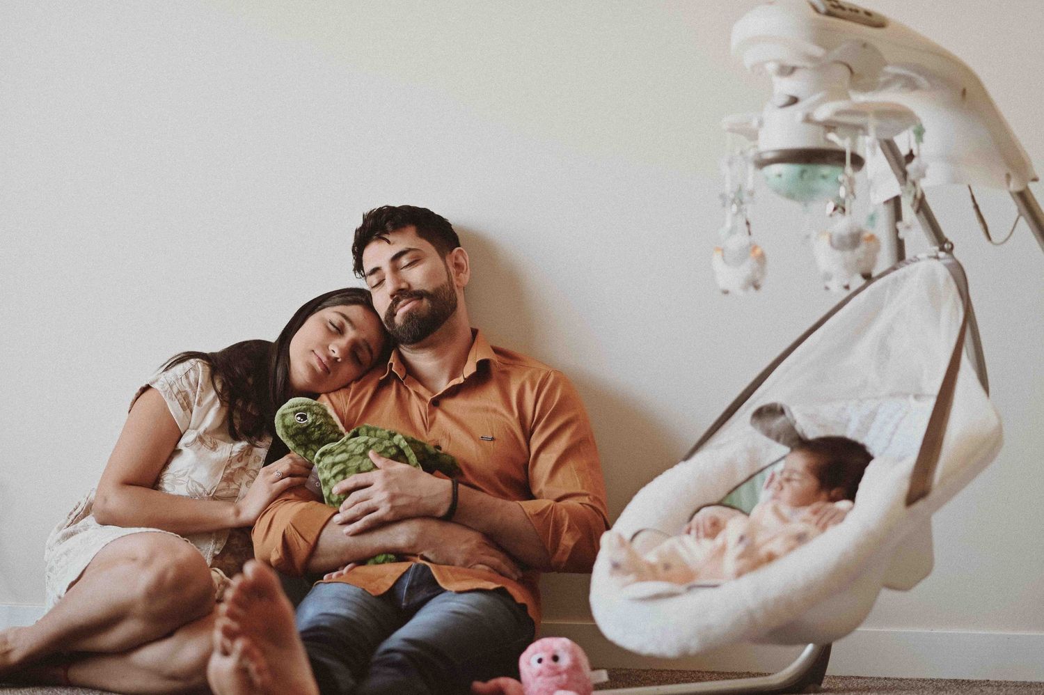 Exhausted new parents lean on each other holding a stuffed toy beside baby in a swing.