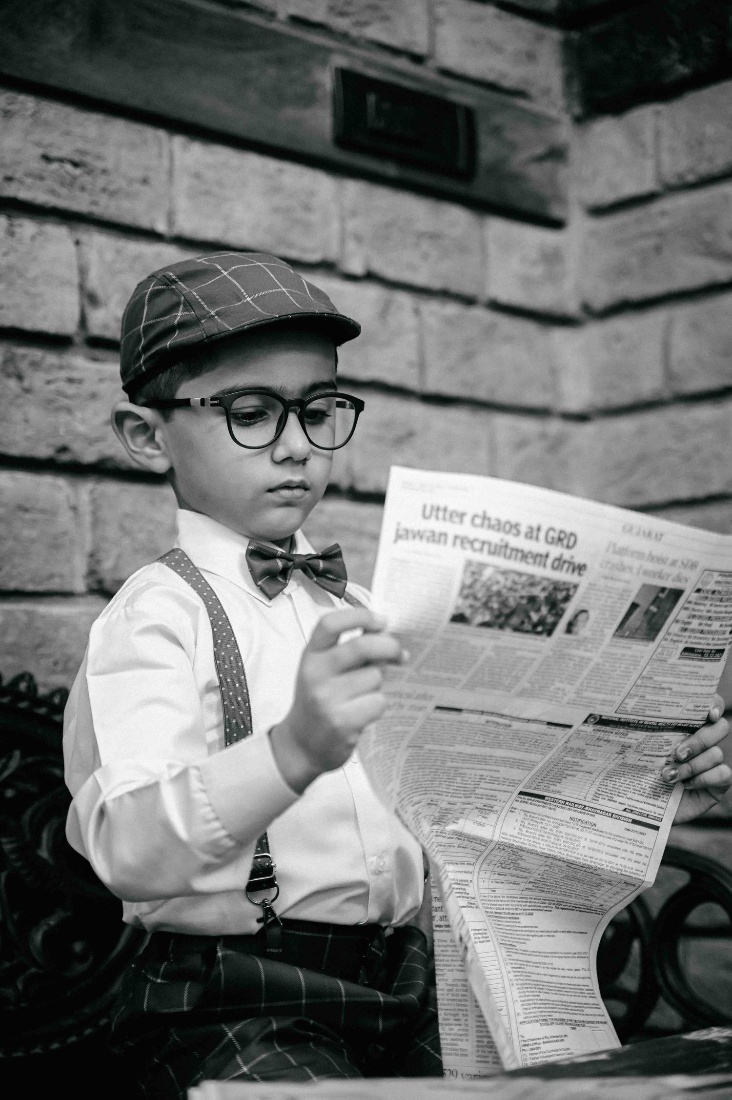 A young boy dressed in vintage style with suspenders, bow tie, cap, and glasses reads a newspaper.