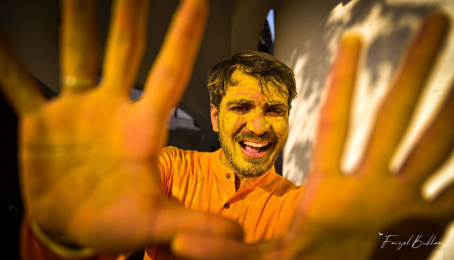 Joyful man covered in yellow holi powder smiling and showing hands to camera at festive celebration.