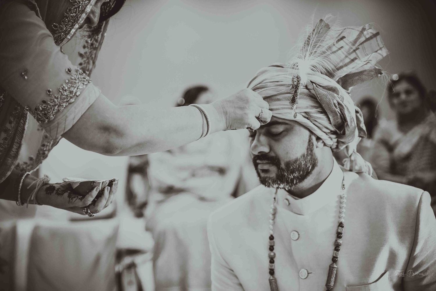 Black and white photo of groom receiving turban during traditional Indian wedding ceremony ritual.