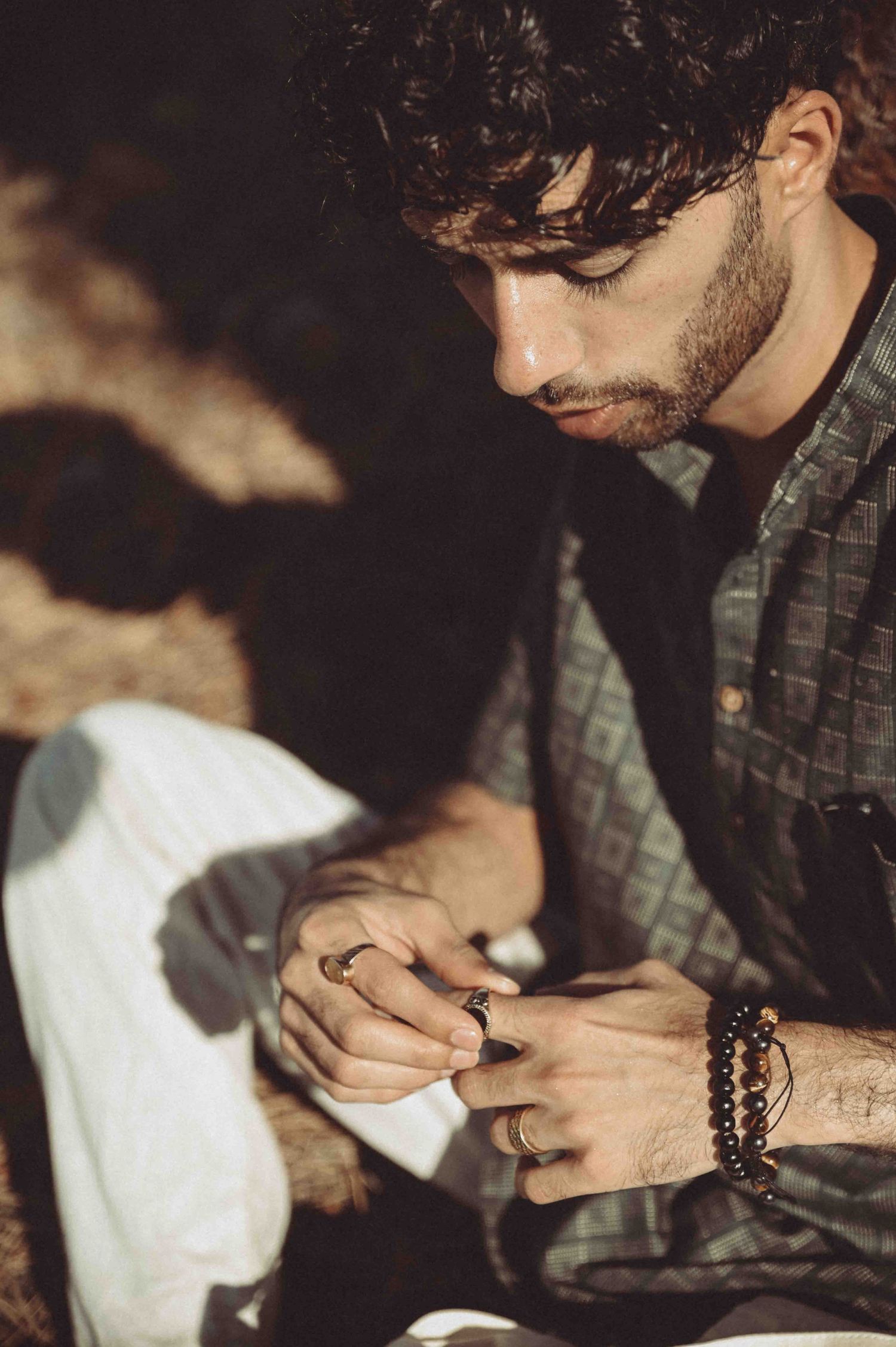 A bearded man with curly hair and beaded bracelet examines jewelry in dappled sunlight.