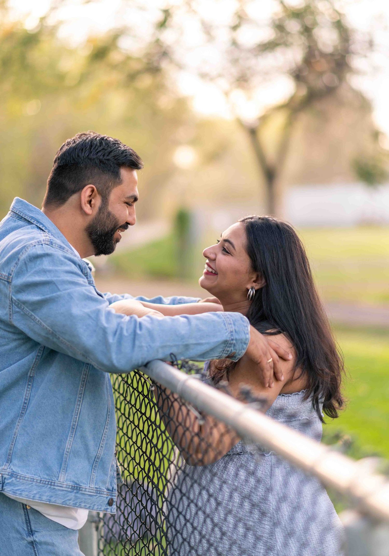 Couple smiling at each other leaning on chain-link fence during golden hour.