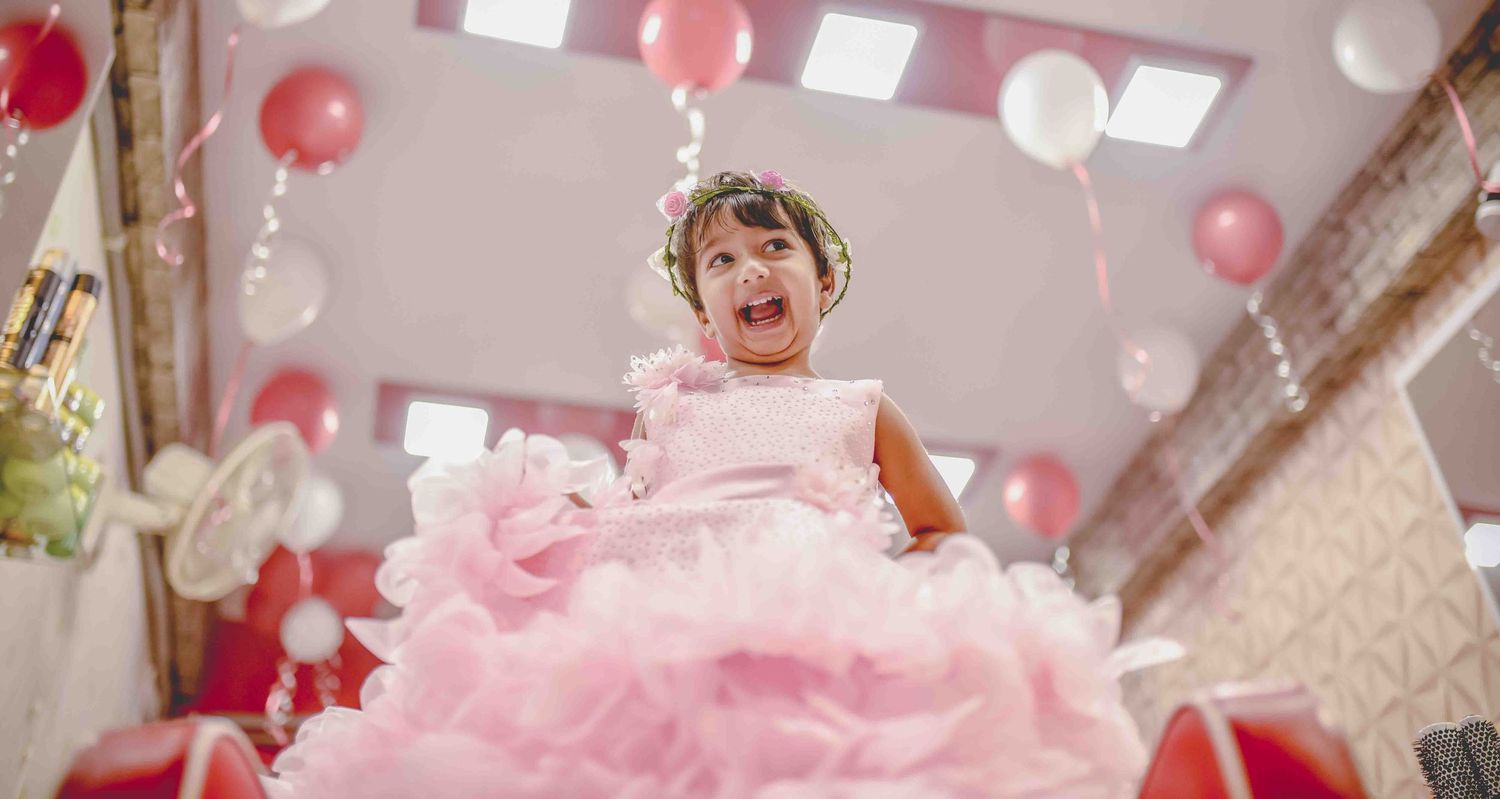 Joyful little girl in pink ball gown laughing under colorful balloons at a birthday party.