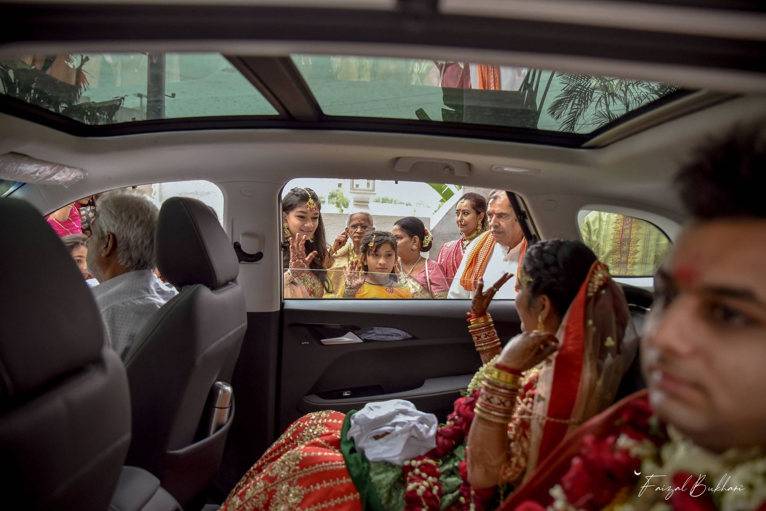 Indian bride in red bridal attire sitting in car while family members gather outside vehicle window.