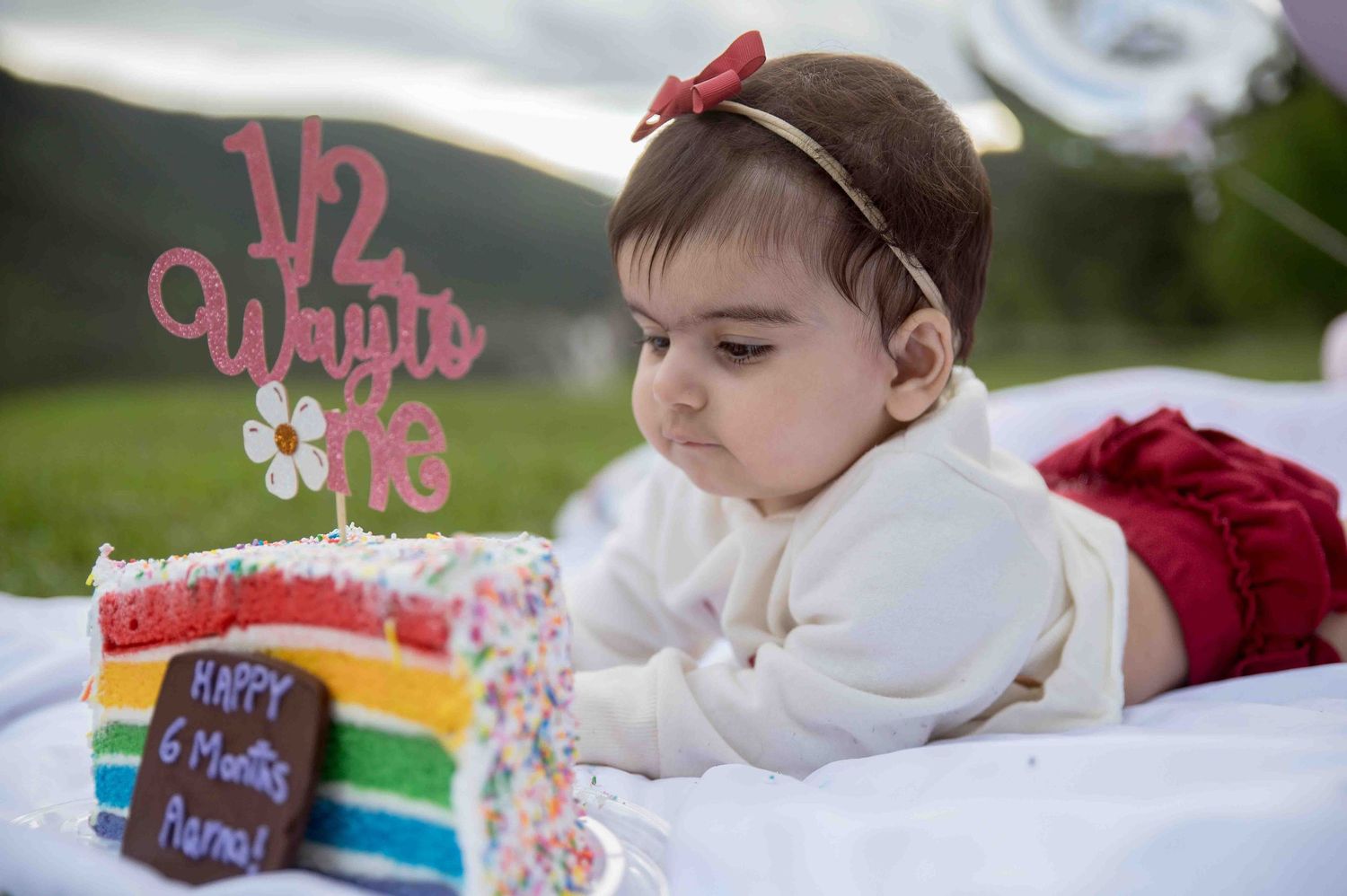Baby girl in red bow headband gazing at rainbow birthday cake with half birthday topper.