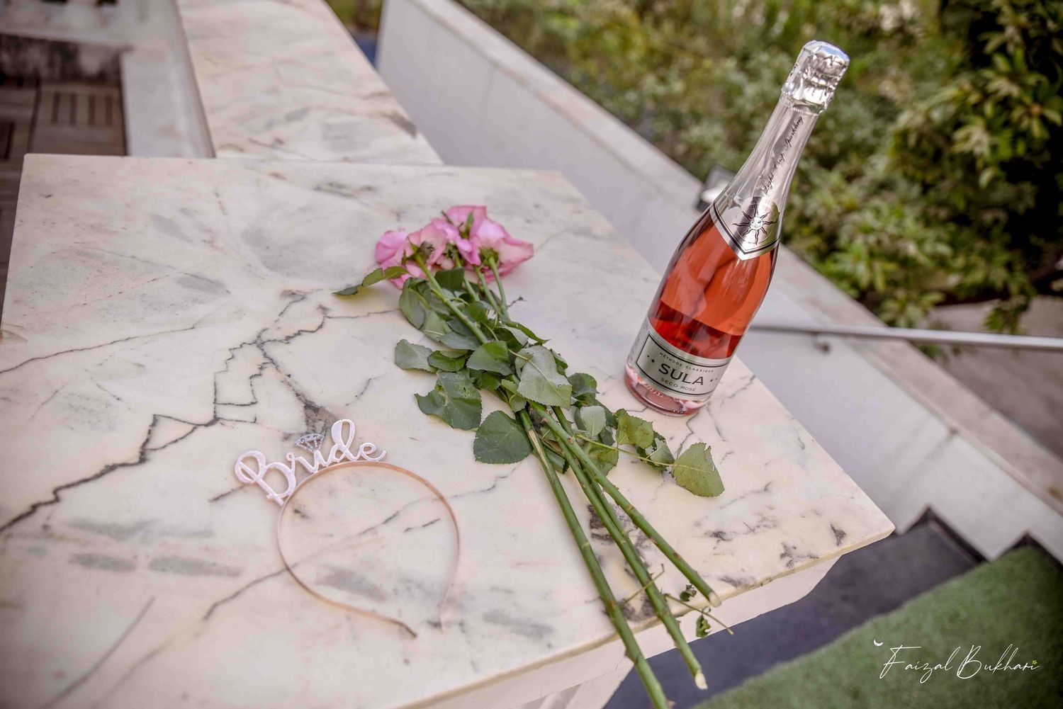 Pink roses, a bride hair hoop, and rose champagne bottle arranged on a marble surface outdoors.
