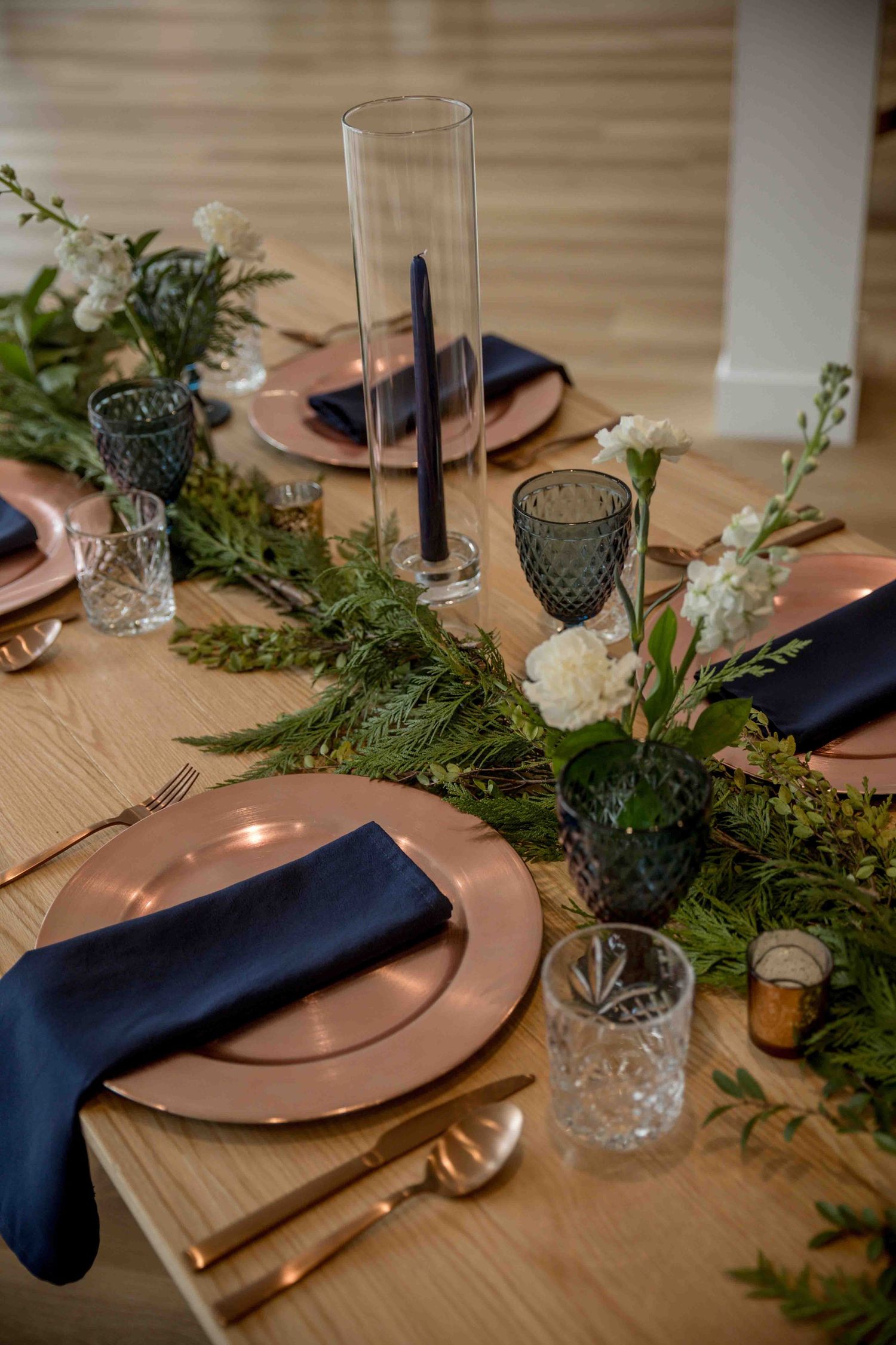 Rustic wedding tablescape with copper charger plates, navy napkins, greenery runner, and dark taper candles.