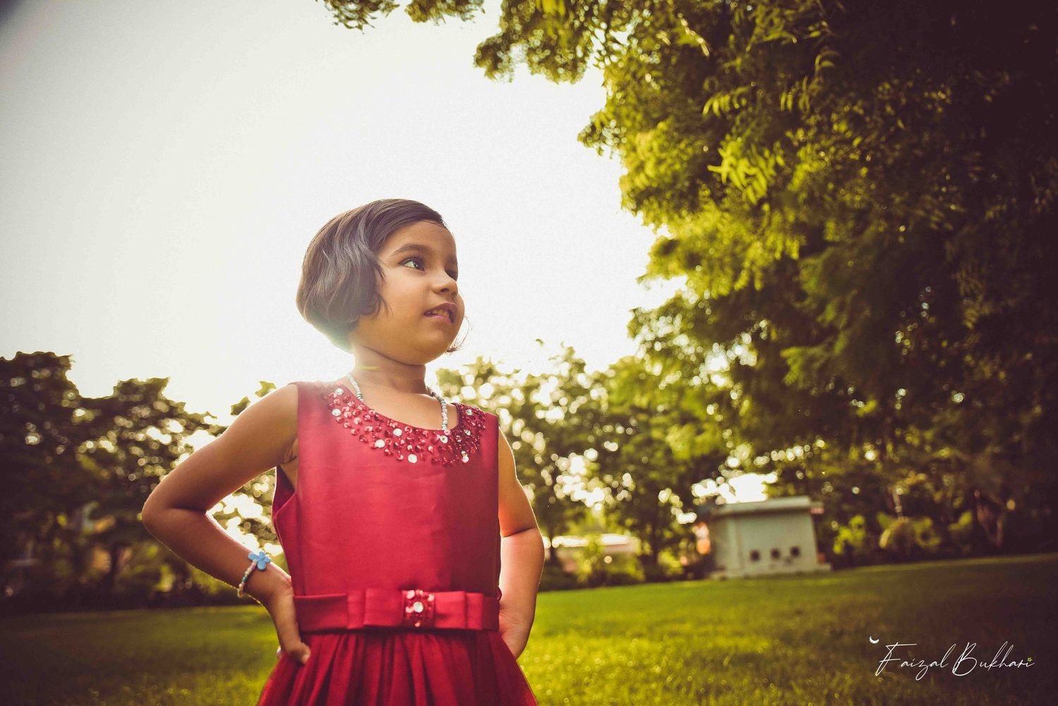 A young girl in a red dress stands confidently on green grass under warm sunlight near trees.