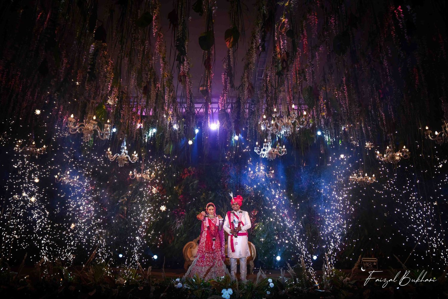 Indian wedding couple standing amid dramatic floral ceiling decor with sparkling fireworks at night celebration.