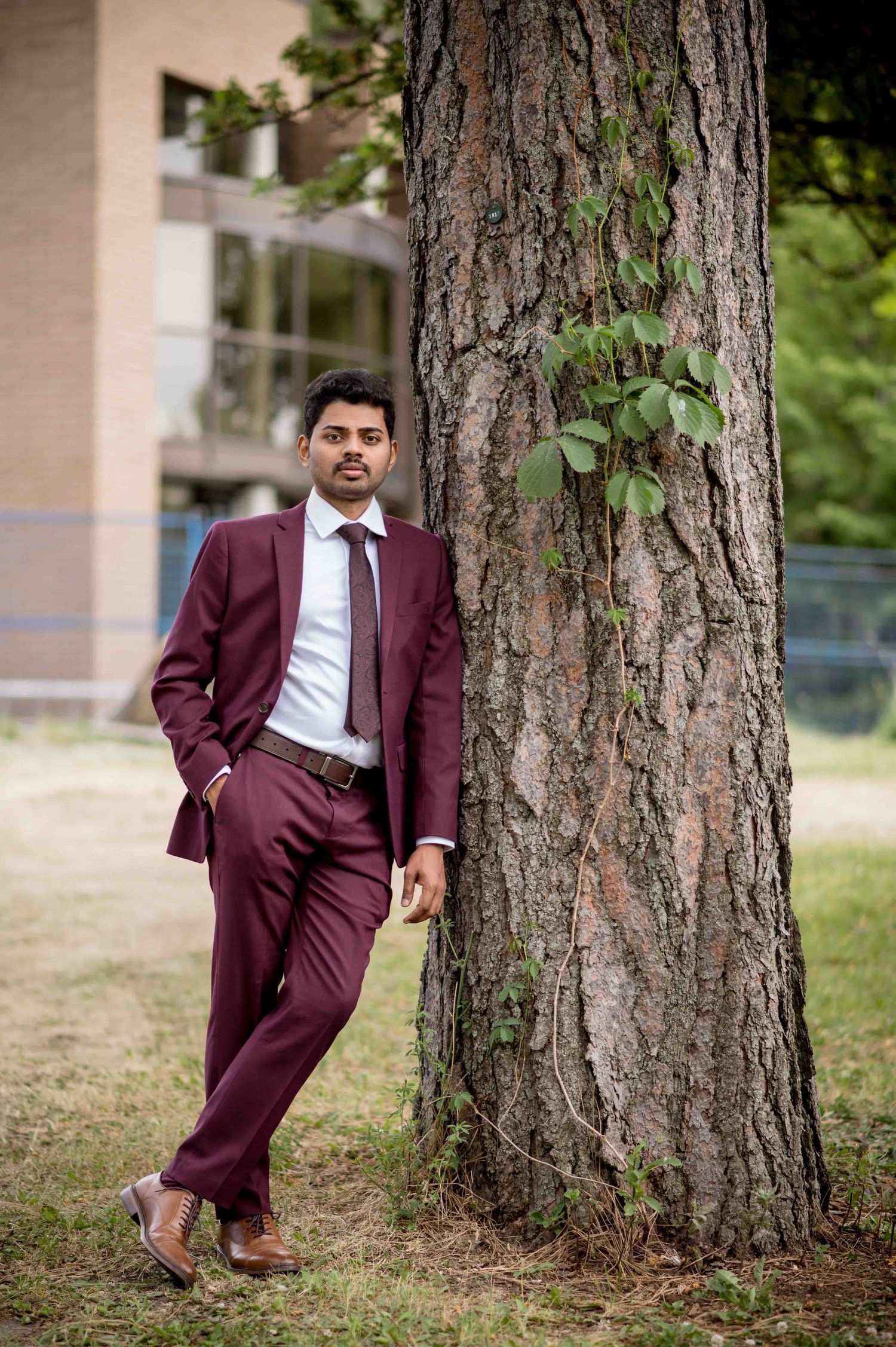 Young man in maroon suit leaning against a large tree trunk on a university campus.