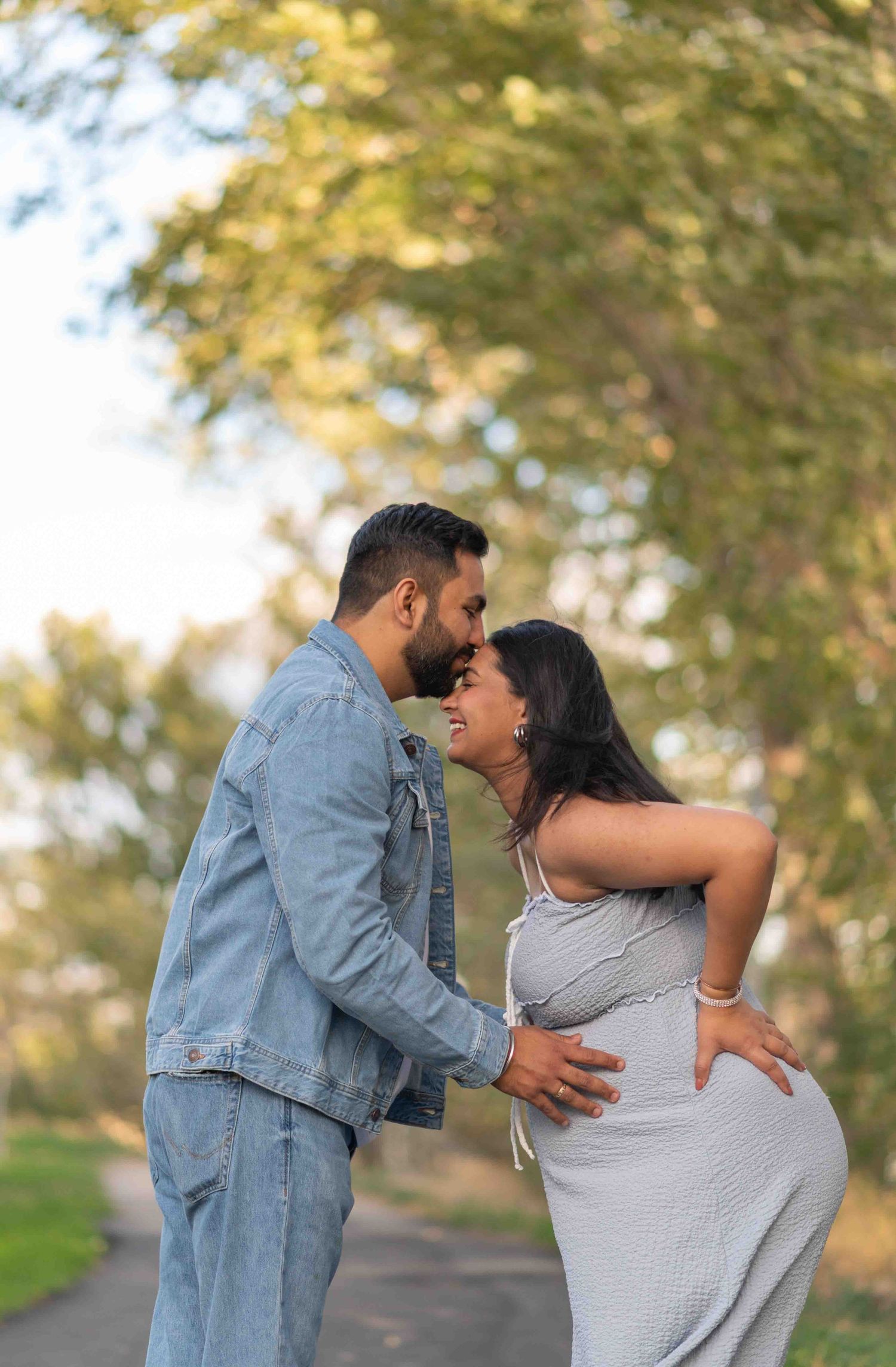 Couple sharing a romantic moment on a tree-lined path, man in denim jacket holding pregnant woman's waist.