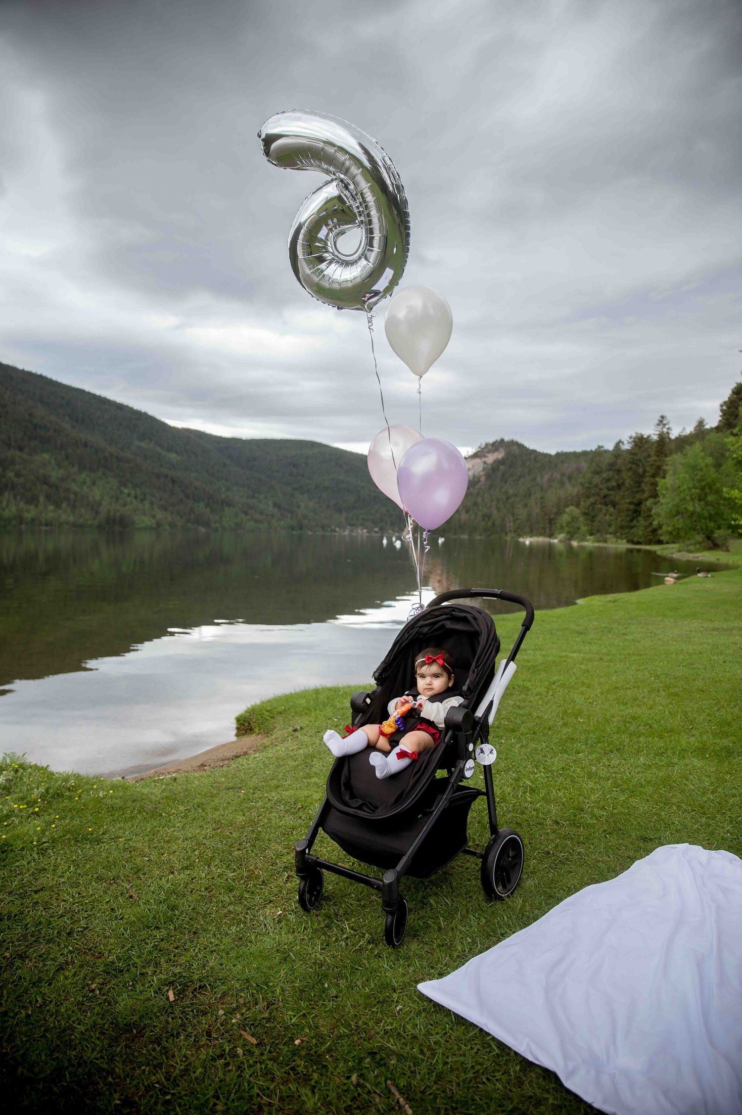 Baby in black stroller with silver number 6 balloon and pastel balloons by scenic mountain lake.