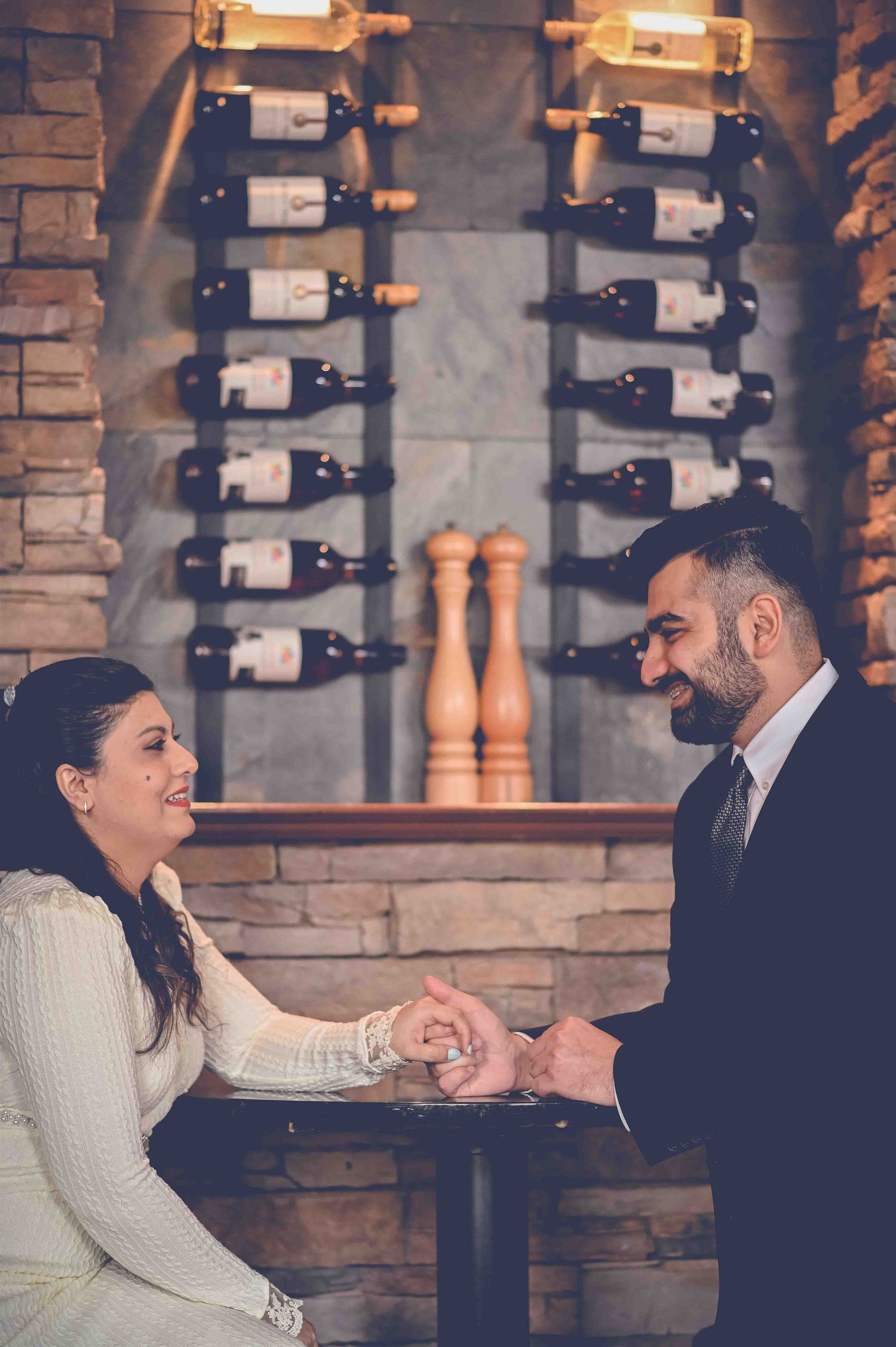 A romantic couple holds hands and smiles at each other in a cozy wine cellar restaurant during their engagement.
