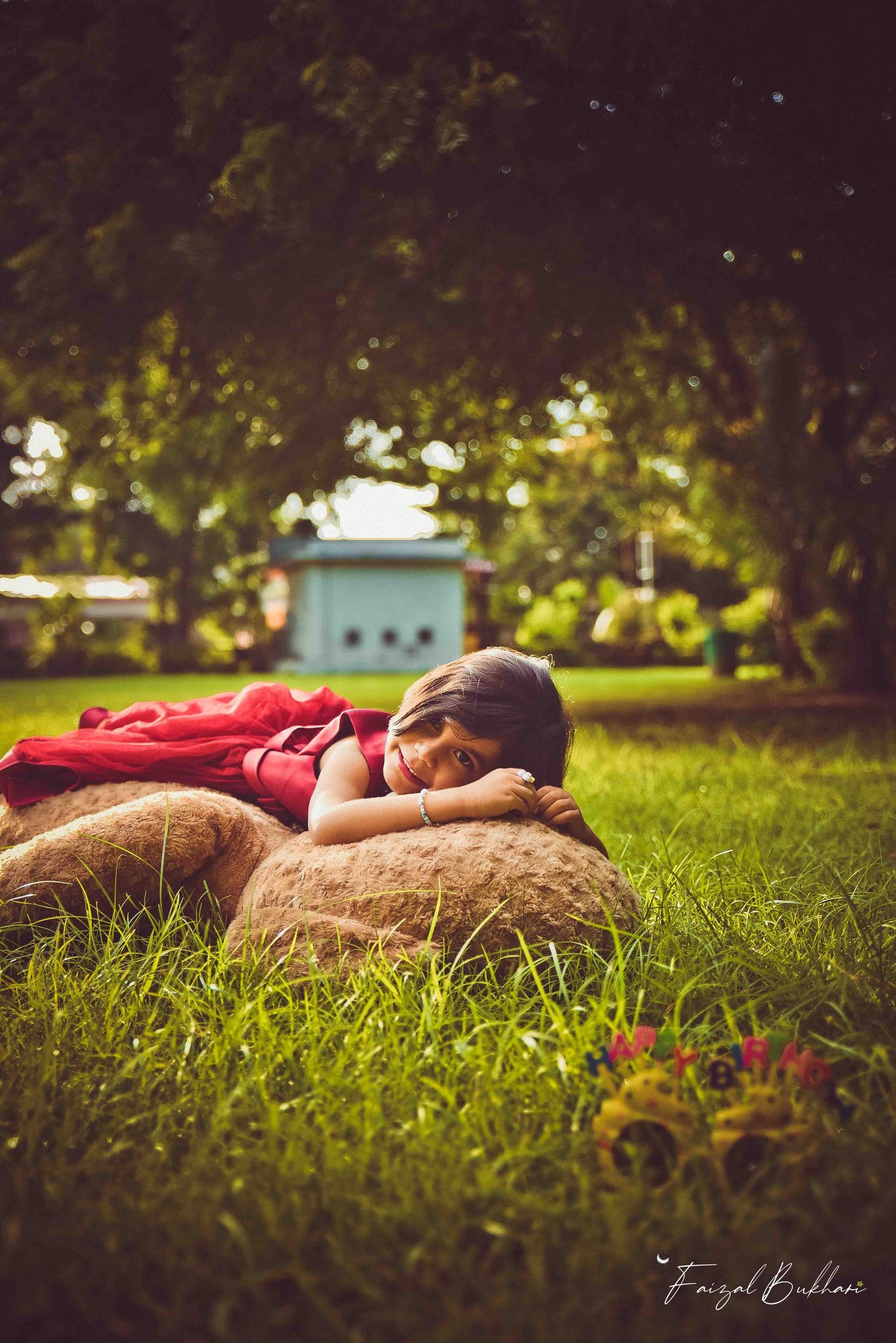 A young girl in a red dress resting peacefully on a giant teddy bear on lush green grass.