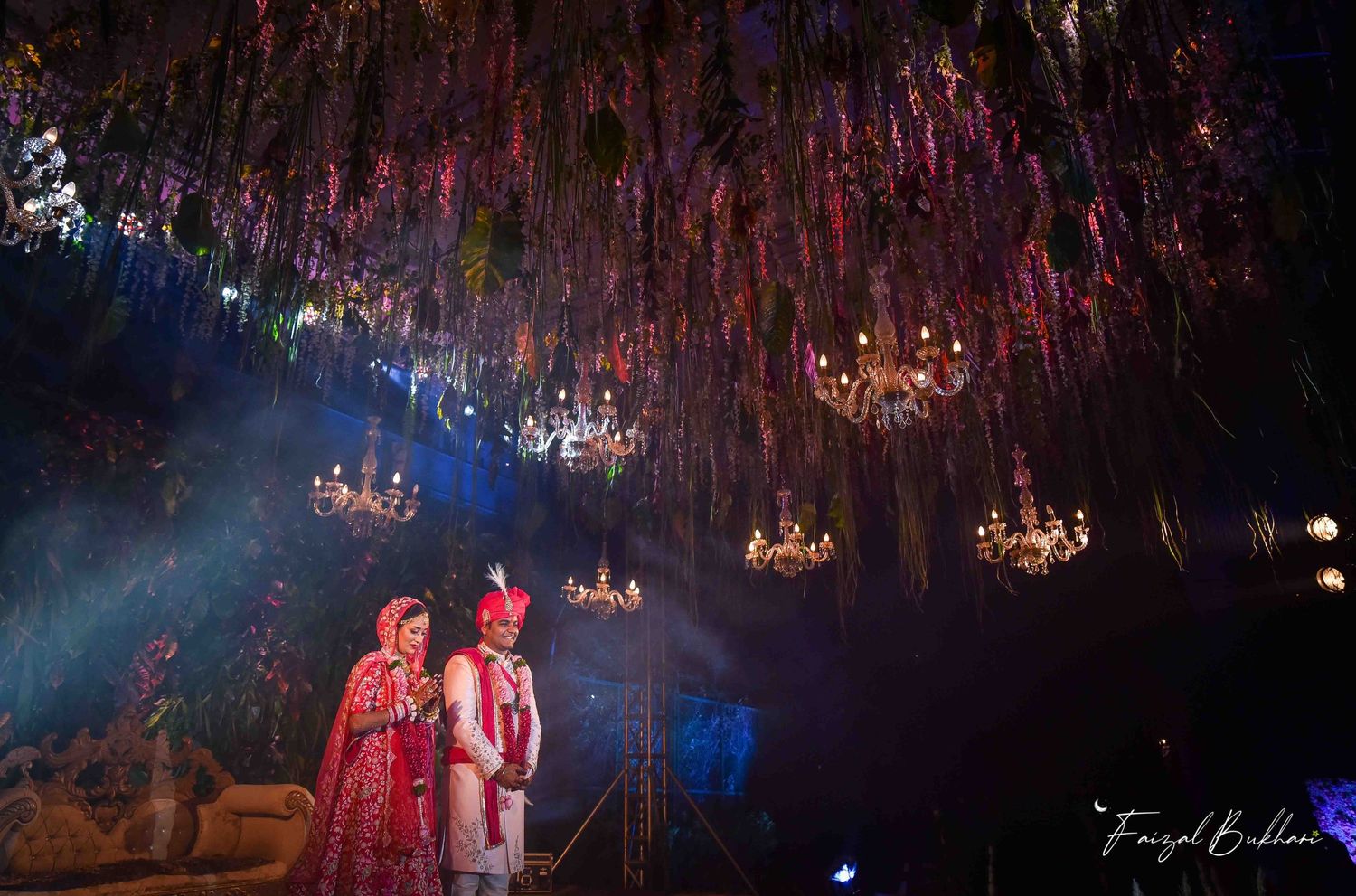 Indian wedding couple posing under stunning floral ceiling installation with crystal chandeliers at nighttime ceremony.