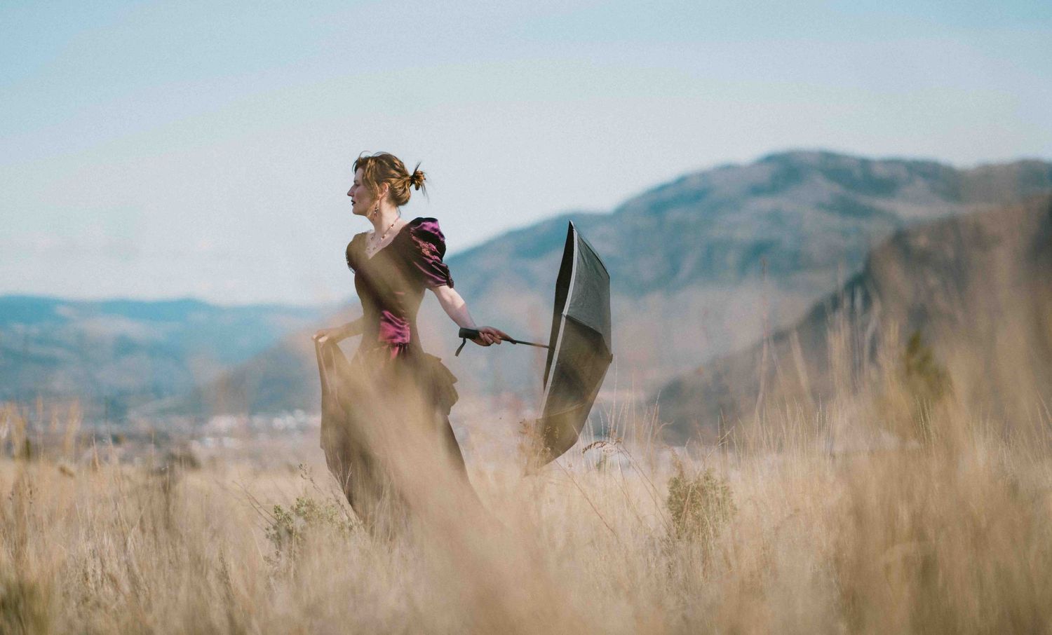 Woman in pink dress holds black umbrella while standing in tall dry grass with mountain backdrop.