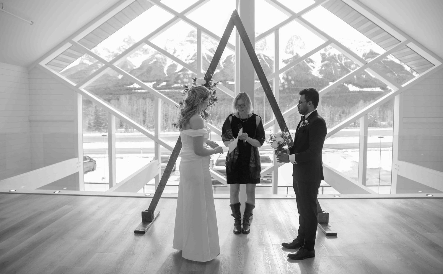 Black and white photo of wedding ceremony with couple exchanging vows beneath a triangular wooden arch.