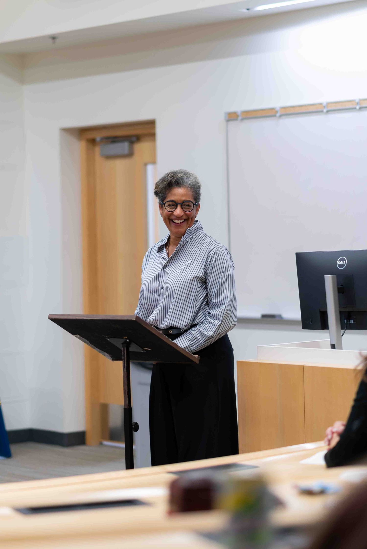 Smiling woman in striped shirt presenting at lecture podium in classroom.