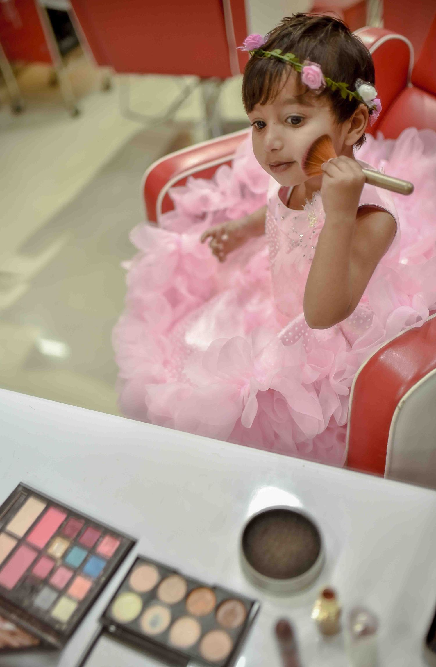 Young girl in pink ruffled dress using a makeup brush at a salon vanity with eyeshadow palette.