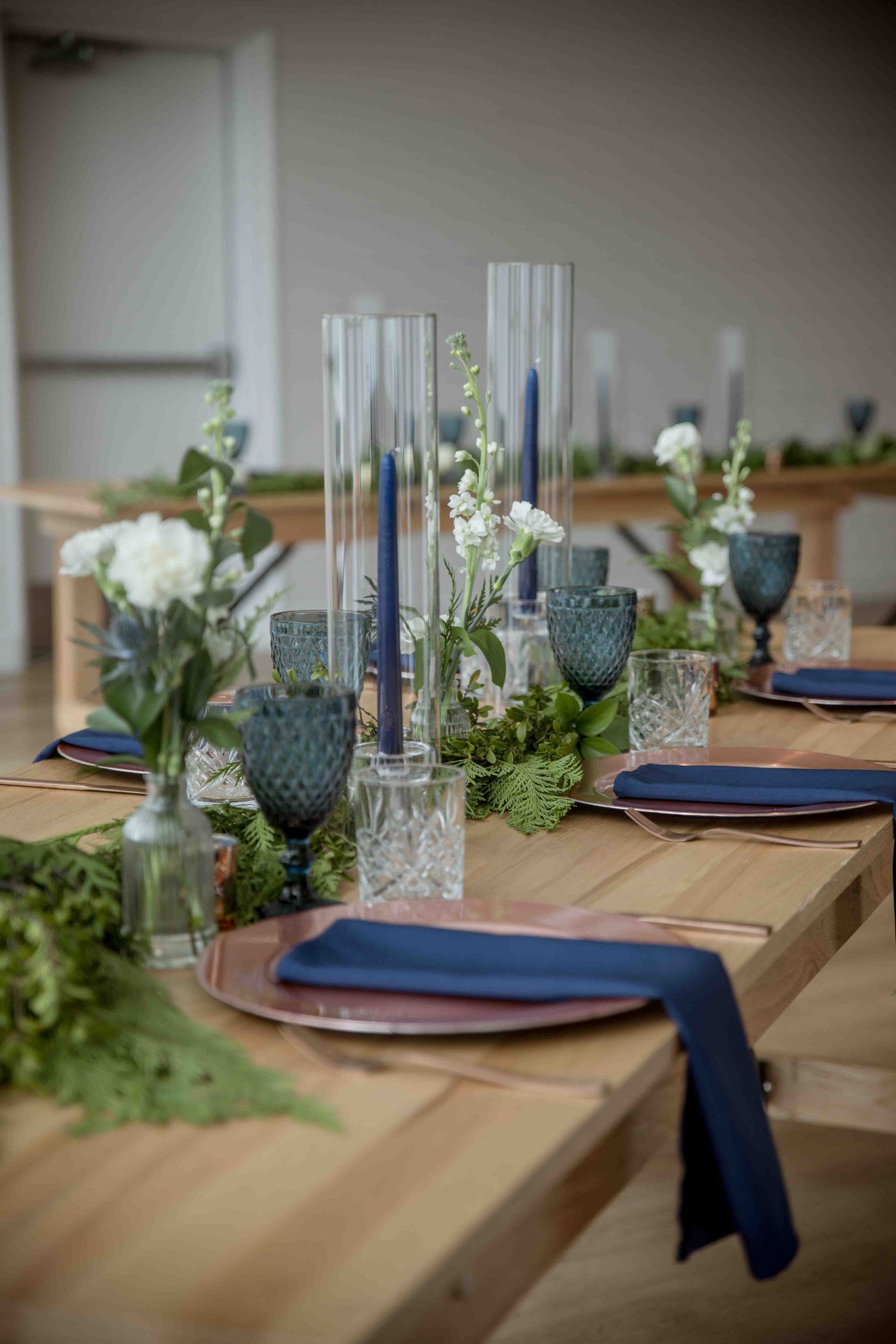 Elegant wedding reception table with navy napkins, blue candles, crystal glasses, and greenery centerpieces.